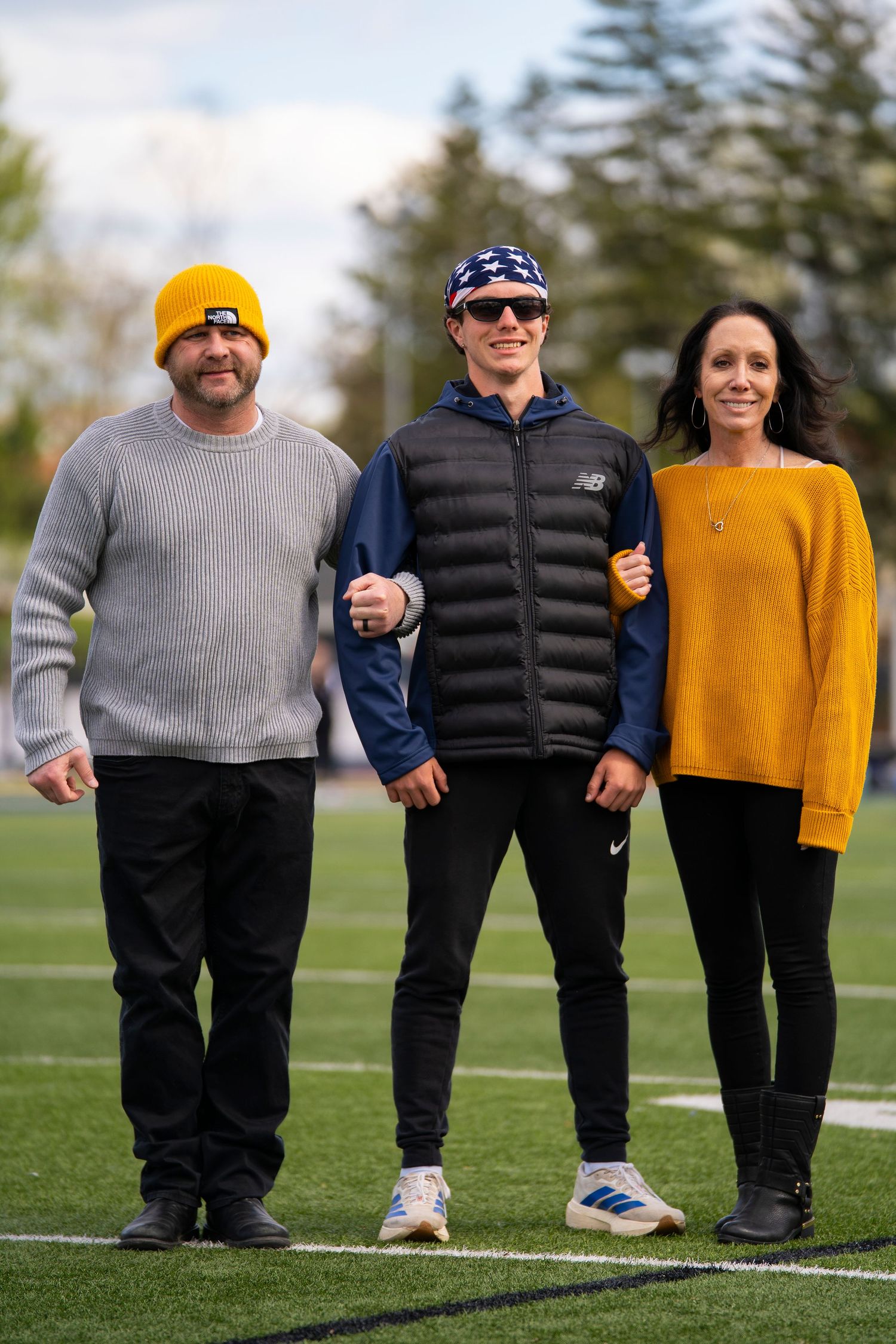 Three people pose together on a football field, wearing casual fall attire including yellow and blue accessories.