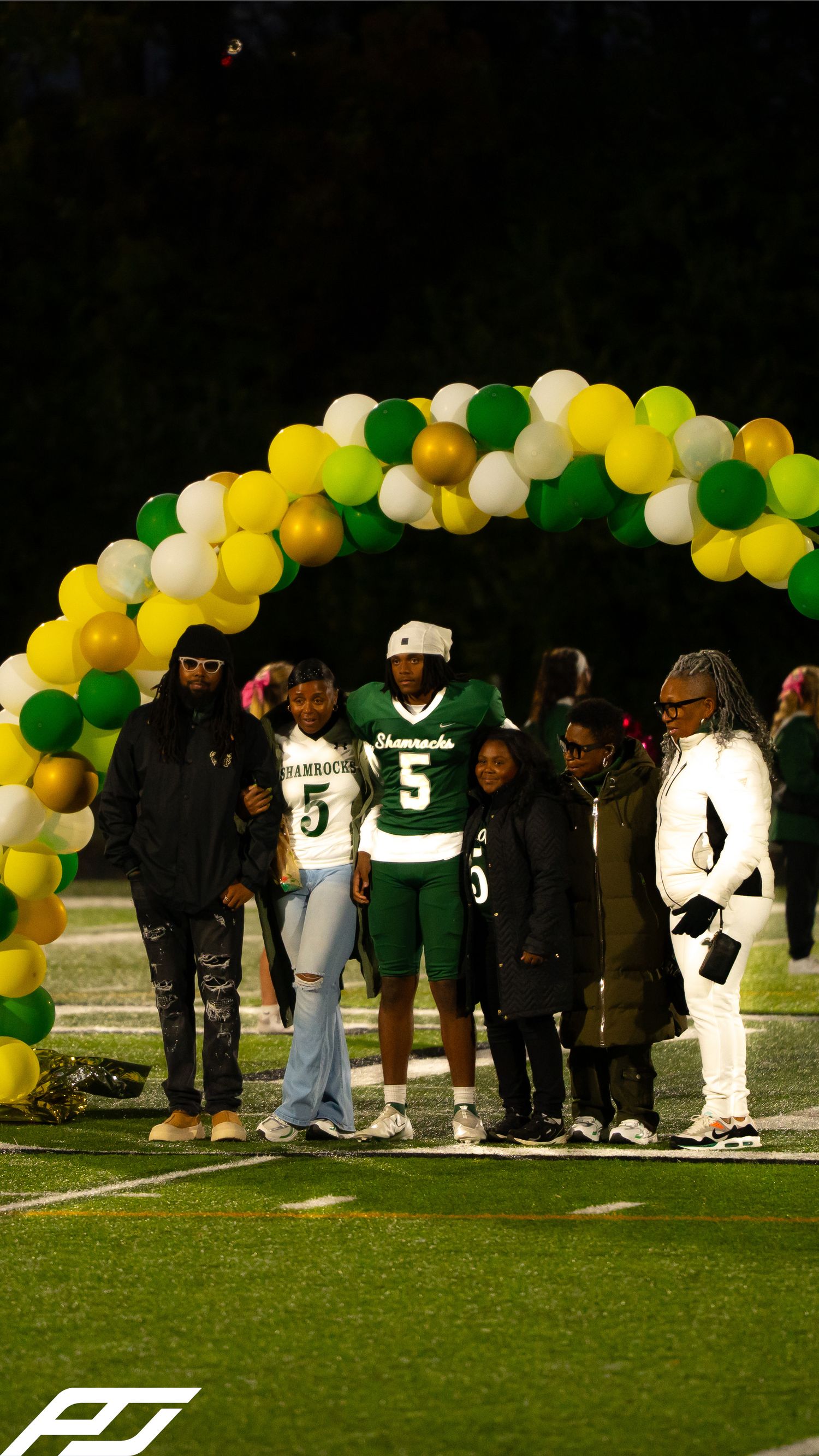 Shamrocks football player number 5 stands with family group under green and yellow balloon arch at night.