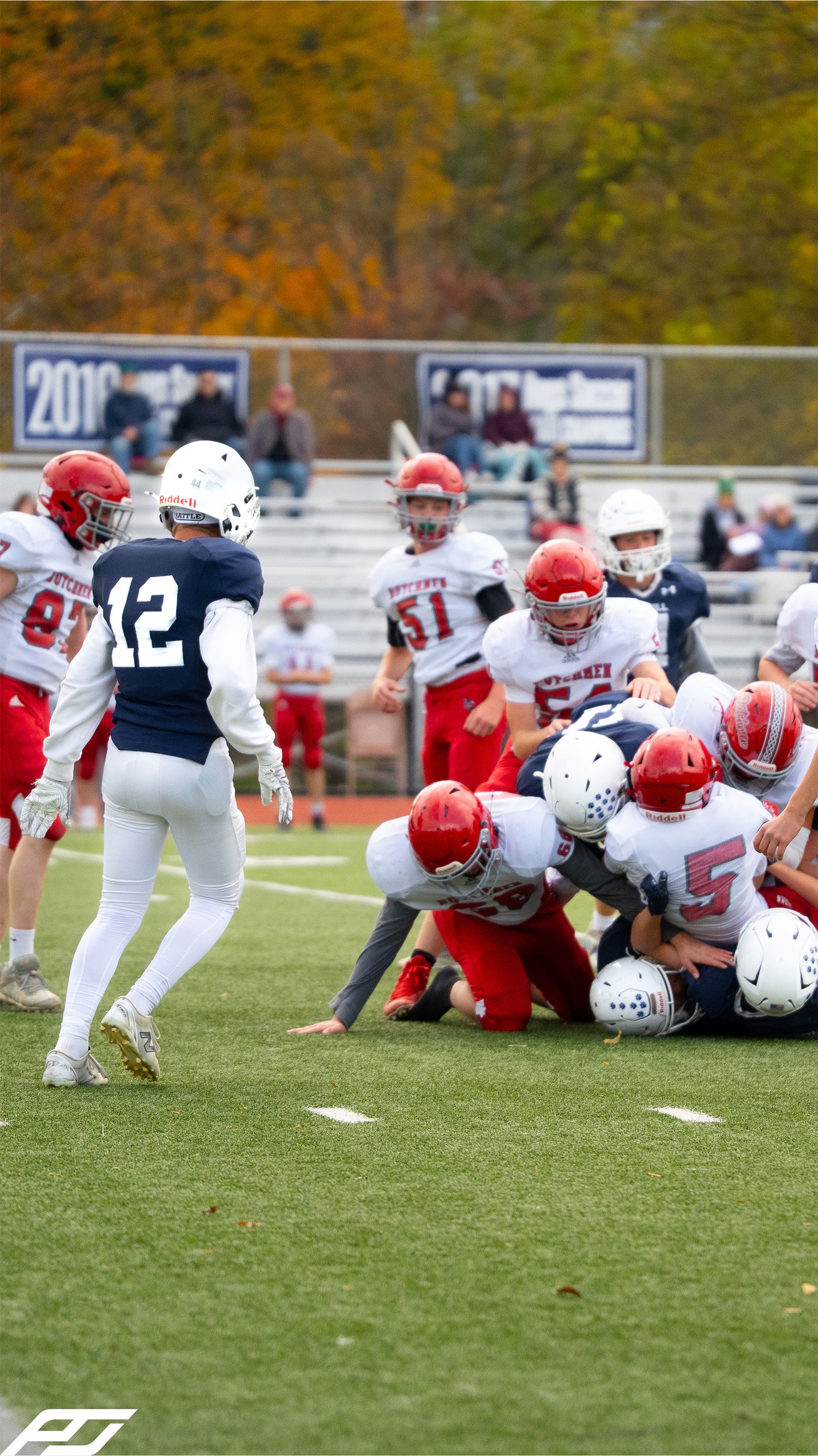 Youth football players in red uniforms tackle a ball carrier while a navy blue player number 12 watches nearby.