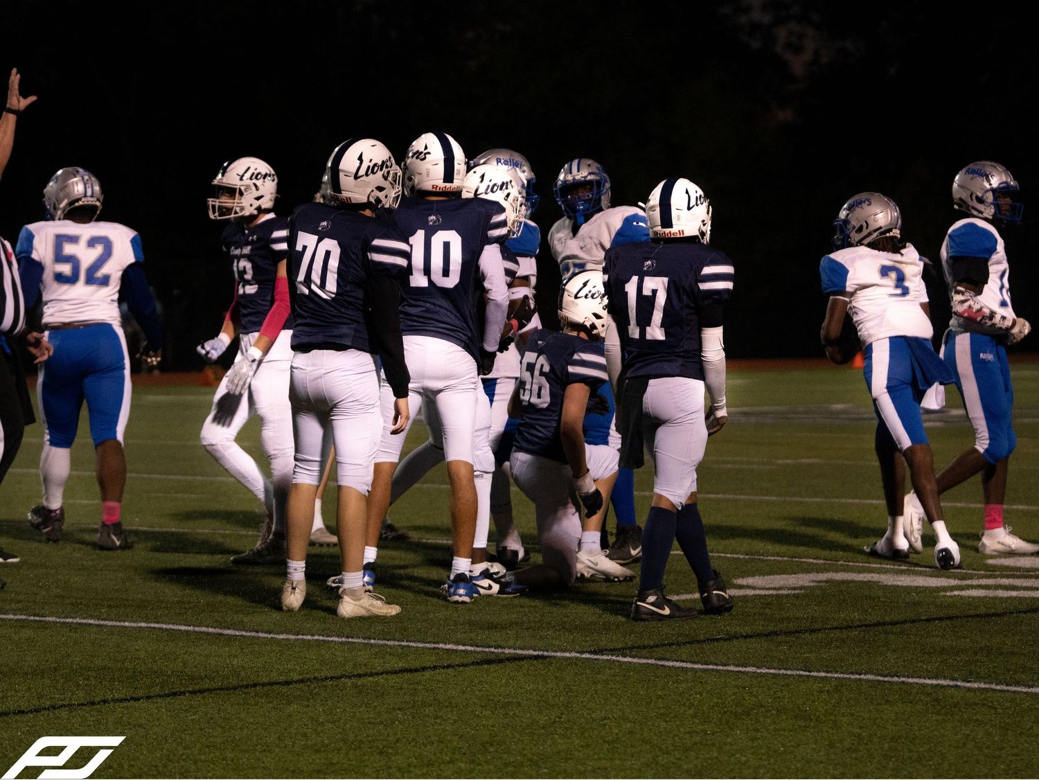 High school football players in navy uniforms numbers 70 and 10 gather on field during night game.