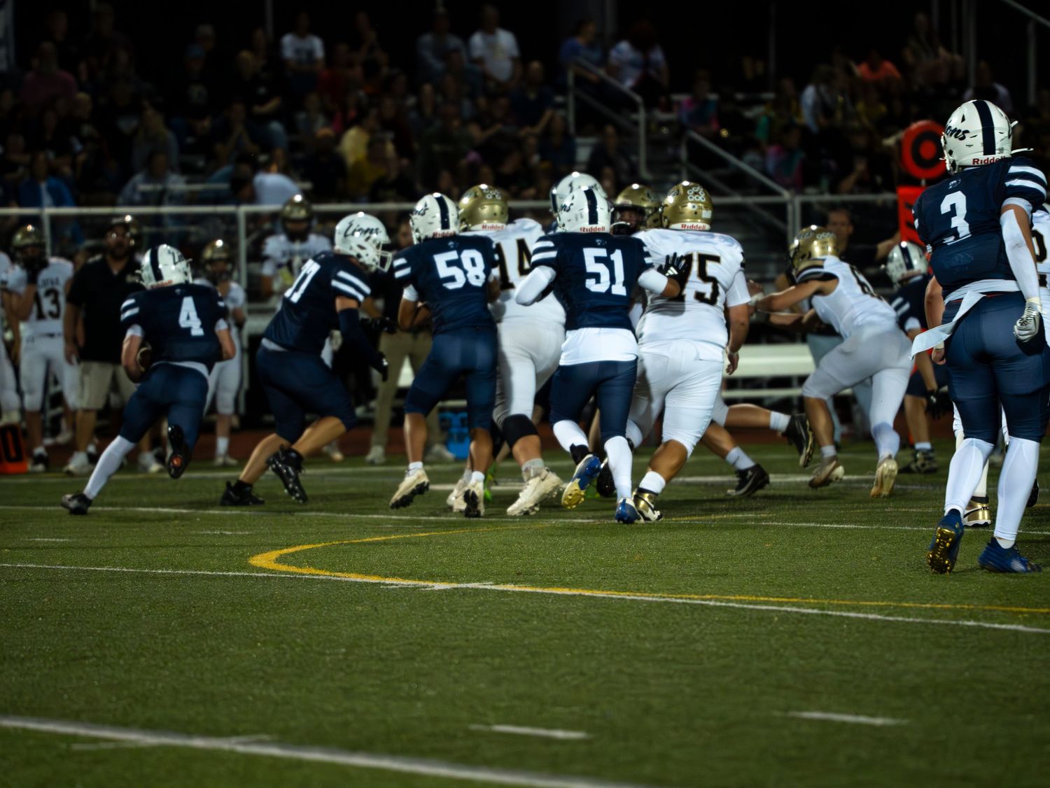 High school football players in blue and white uniforms engage in a line scrimmage during a night game.