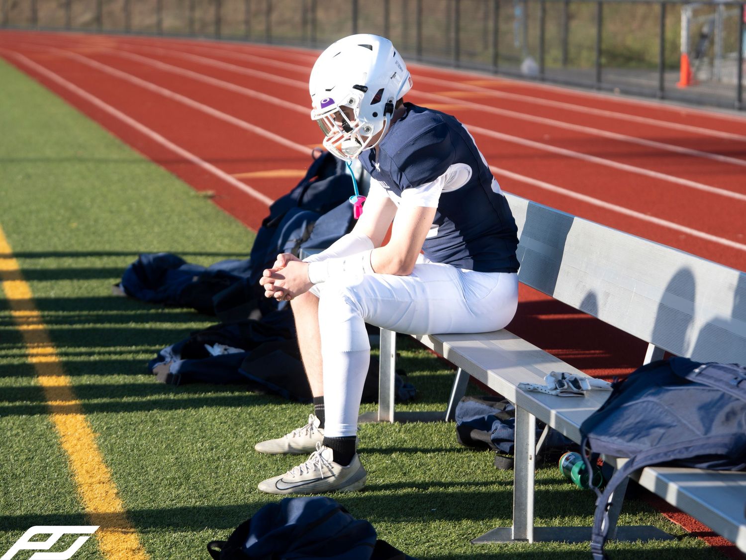 Youth football player in navy uniform and helmet sits alone on sideline bench during game.