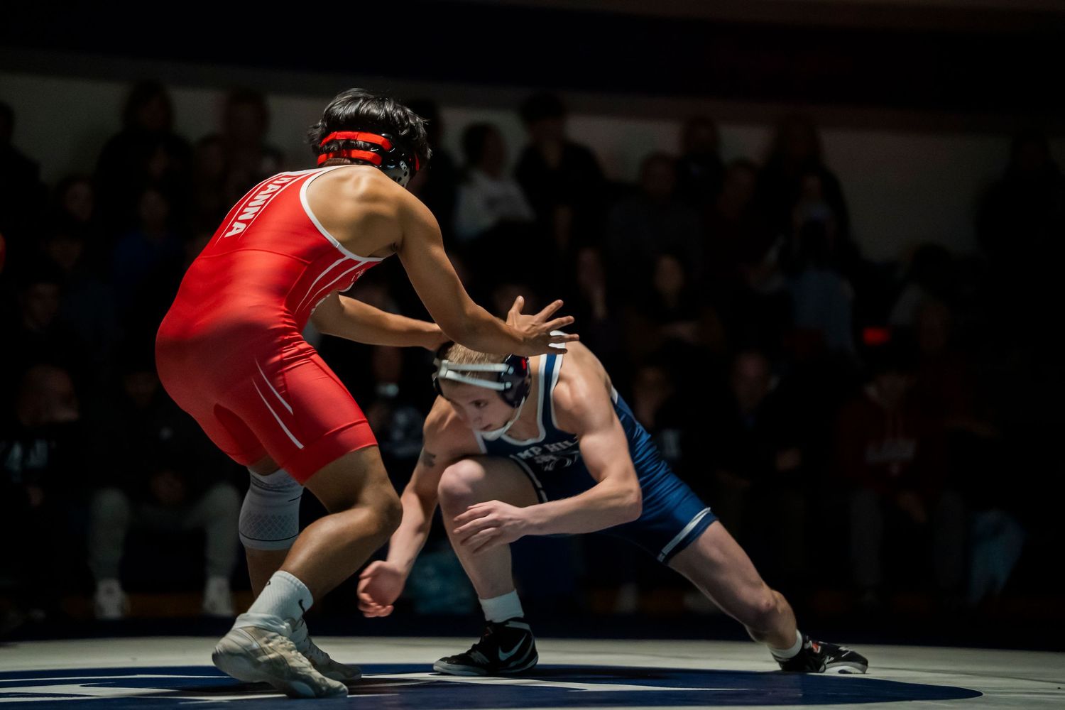 Two wrestlers in red and blue singlets face off in a standing grappling position during a match.