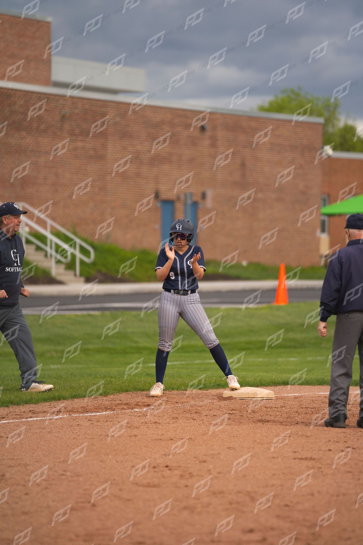 Softball player celebrates on first base during a game, with coaches nearby on a cloudy day.