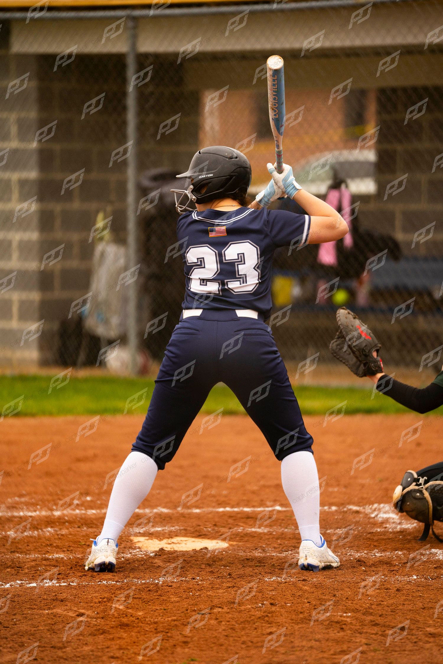 Softball player #23 in navy uniform stands at home plate in batting stance ready to hit on a dirt field.