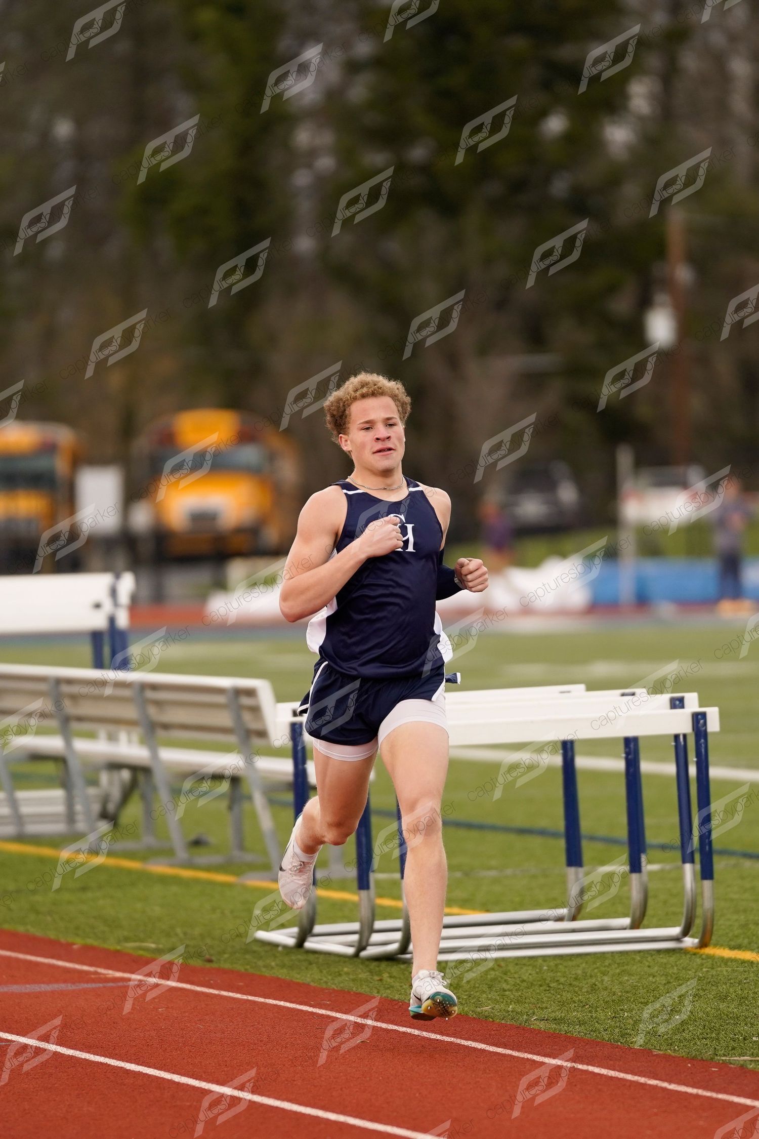 Male runner in navy uniform competing on a red track with hurdles in the background.