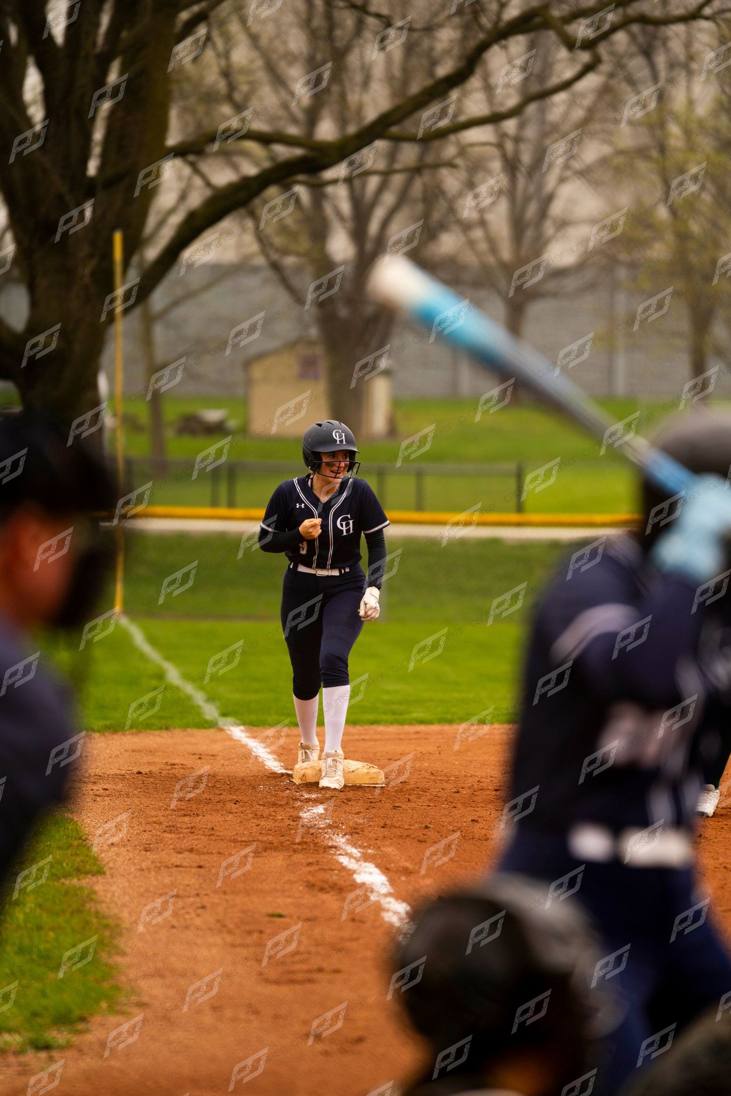 Softball player in navy uniform stands on base smiling as a batter swings in the foreground.