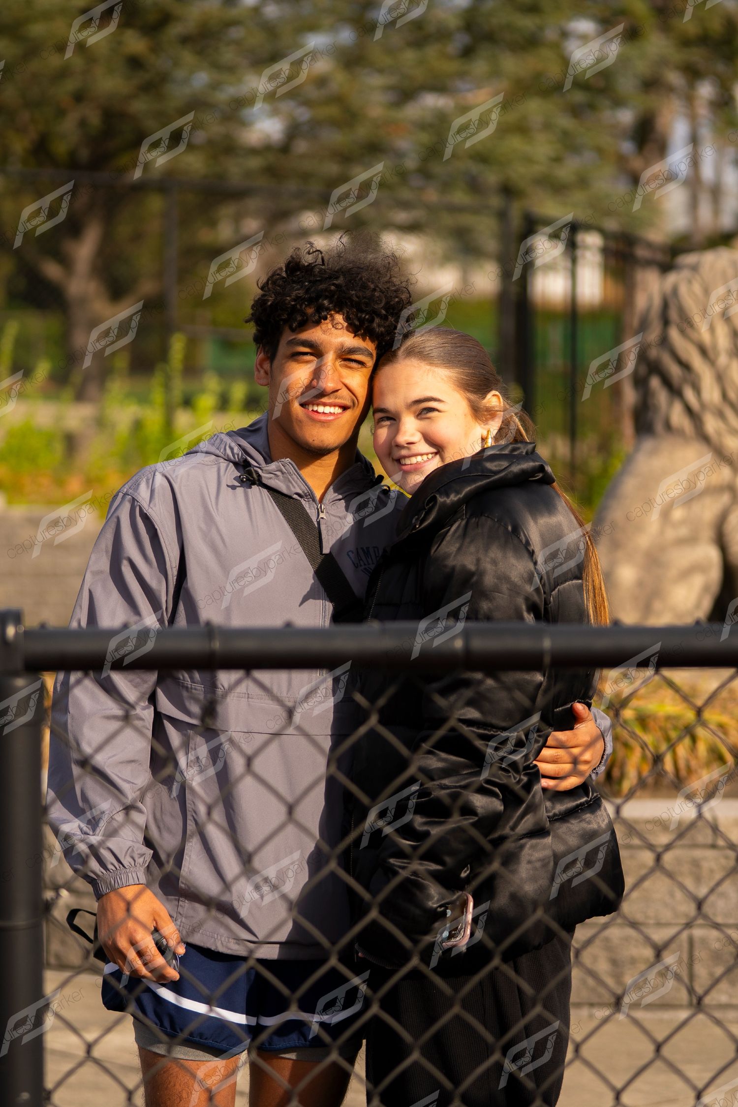 Smiling teenage couple embracing near chain-link fence at outdoor track and field meet.
