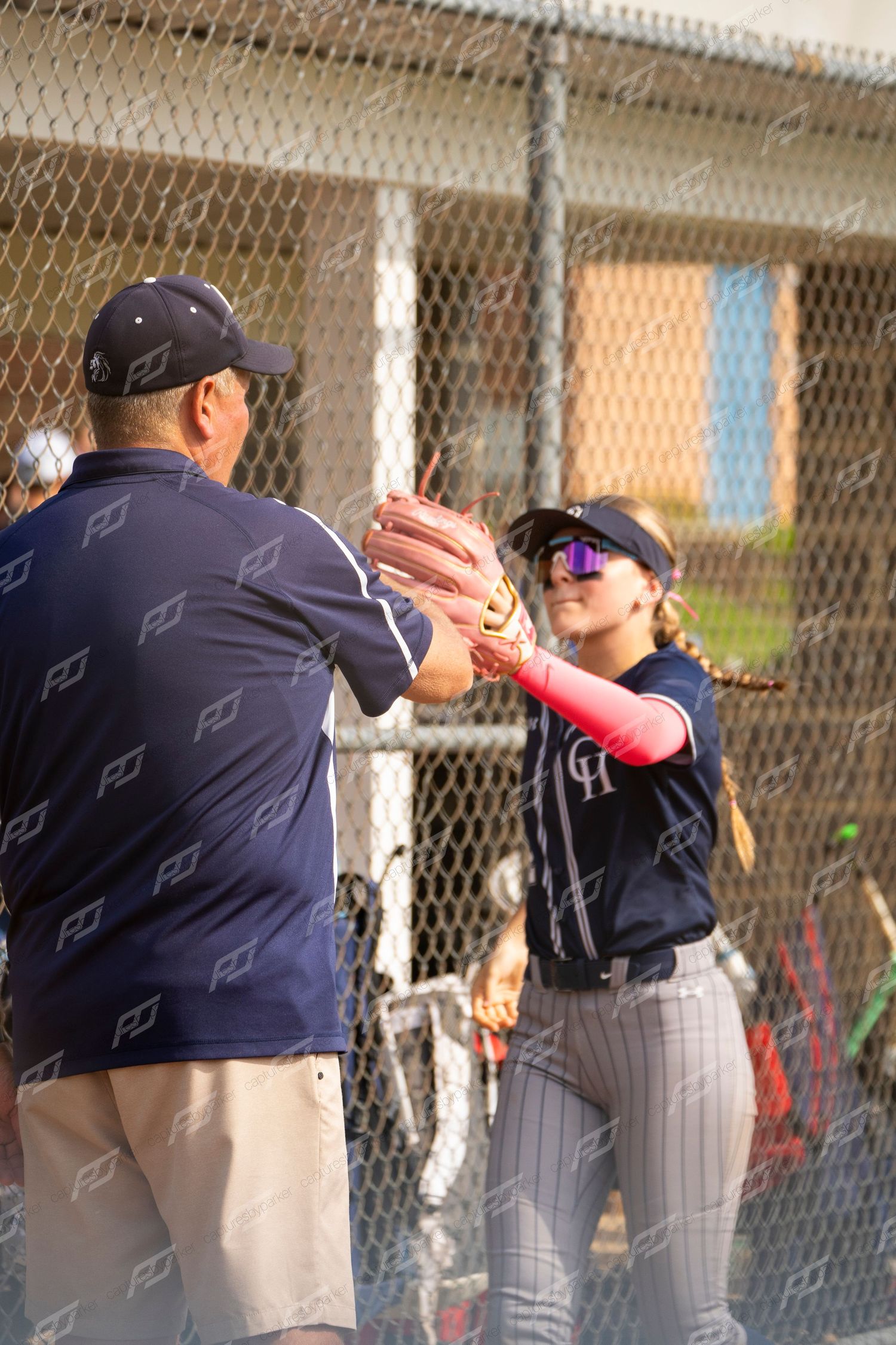 Female softball player high-fives a coach in navy uniform near a chain-link fence.