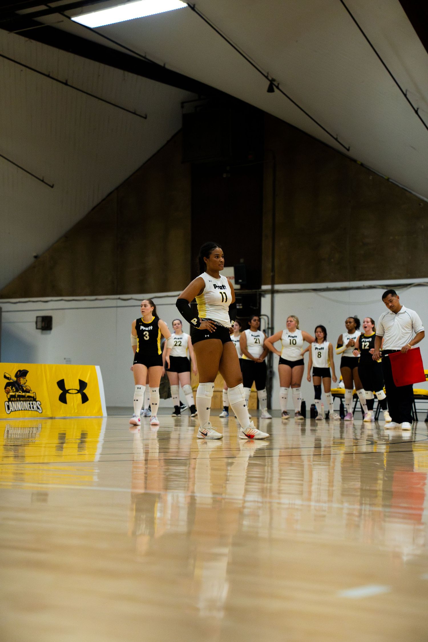 Female volleyball players in black and white uniforms stand on an indoor court during a game warm-up session.