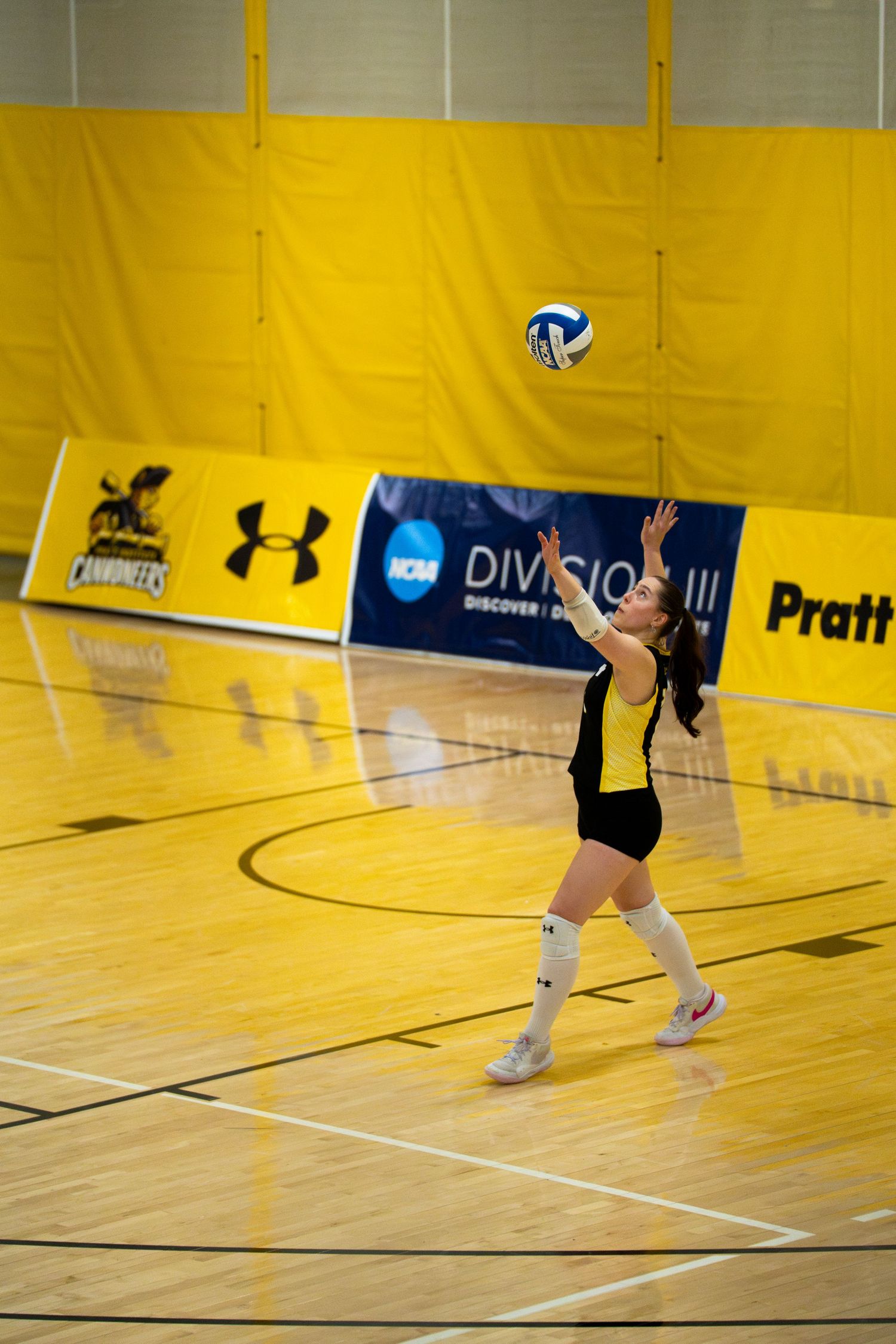 Female volleyball player serving the ball indoors at an NCAA Division III match on a hardwood court.