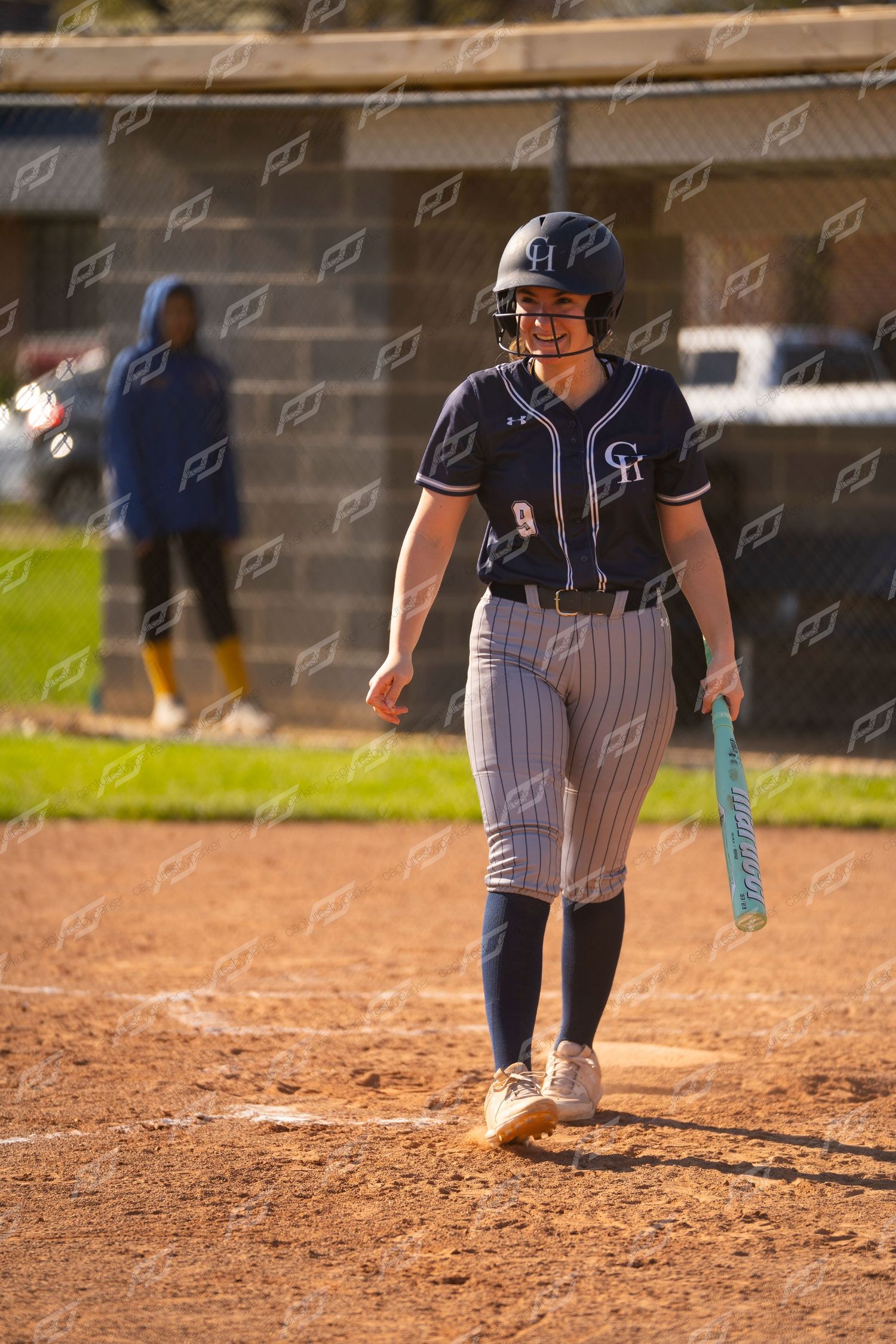 Softball player in navy pinstripe uniform #9 smiling while holding bat at home plate during game.