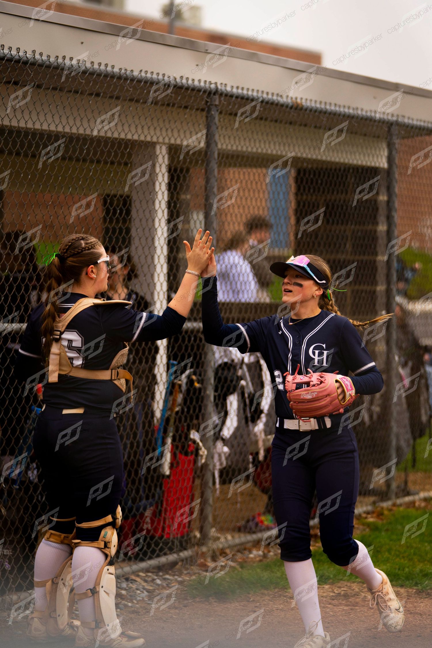 Two softball players in navy uniforms celebrating with a high five near the dugout fence after play.