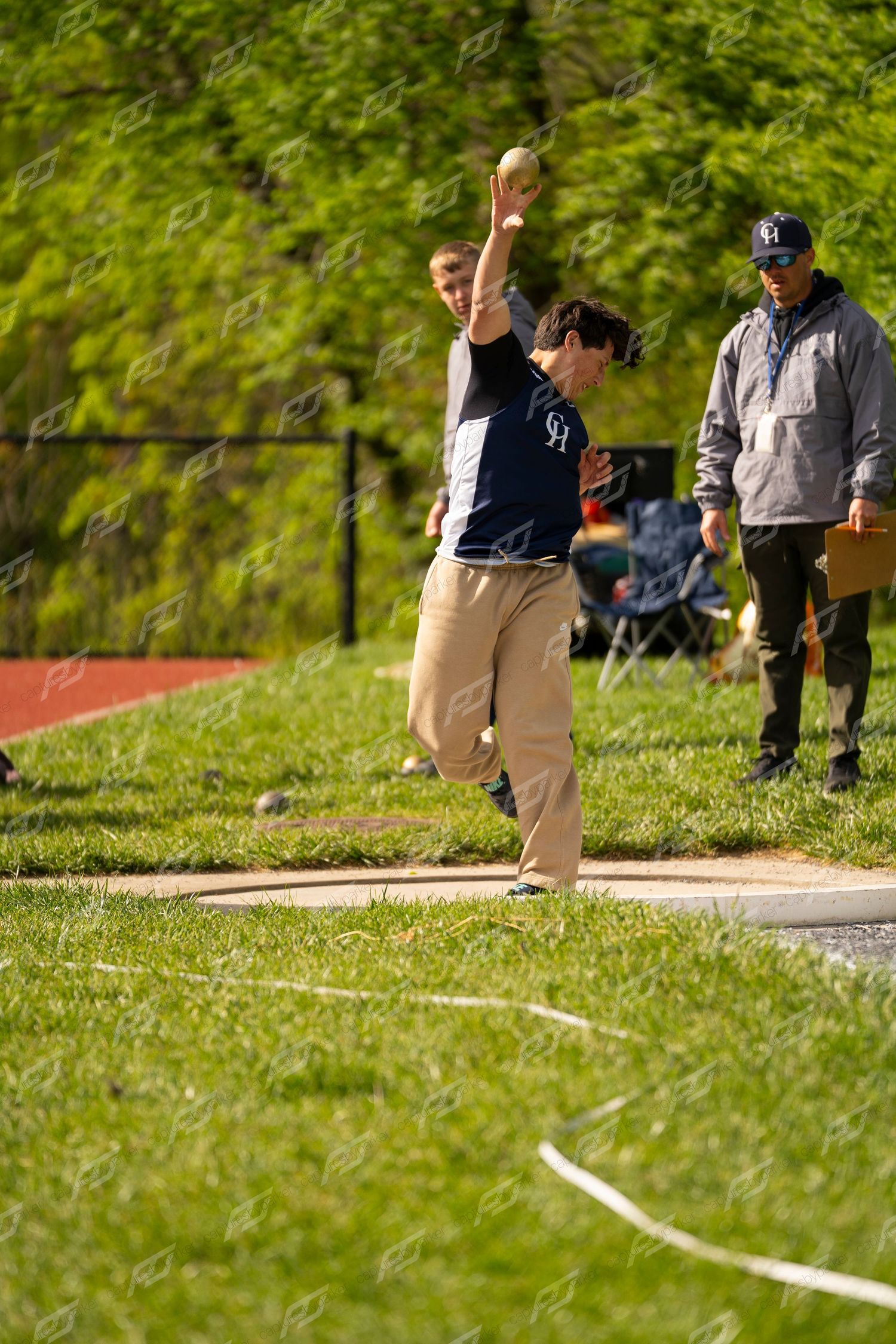 Male athlete in navy CH uniform throwing shot put while official observes at outdoor track meet.