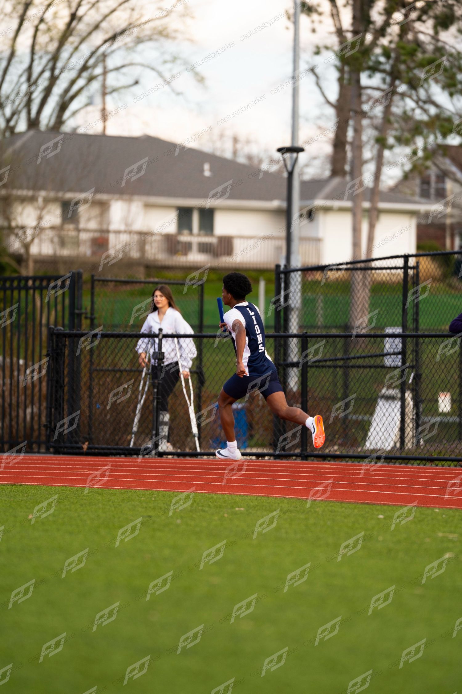 High school male track athlete sprints on red outdoor track during relay race while spectator watches nearby.