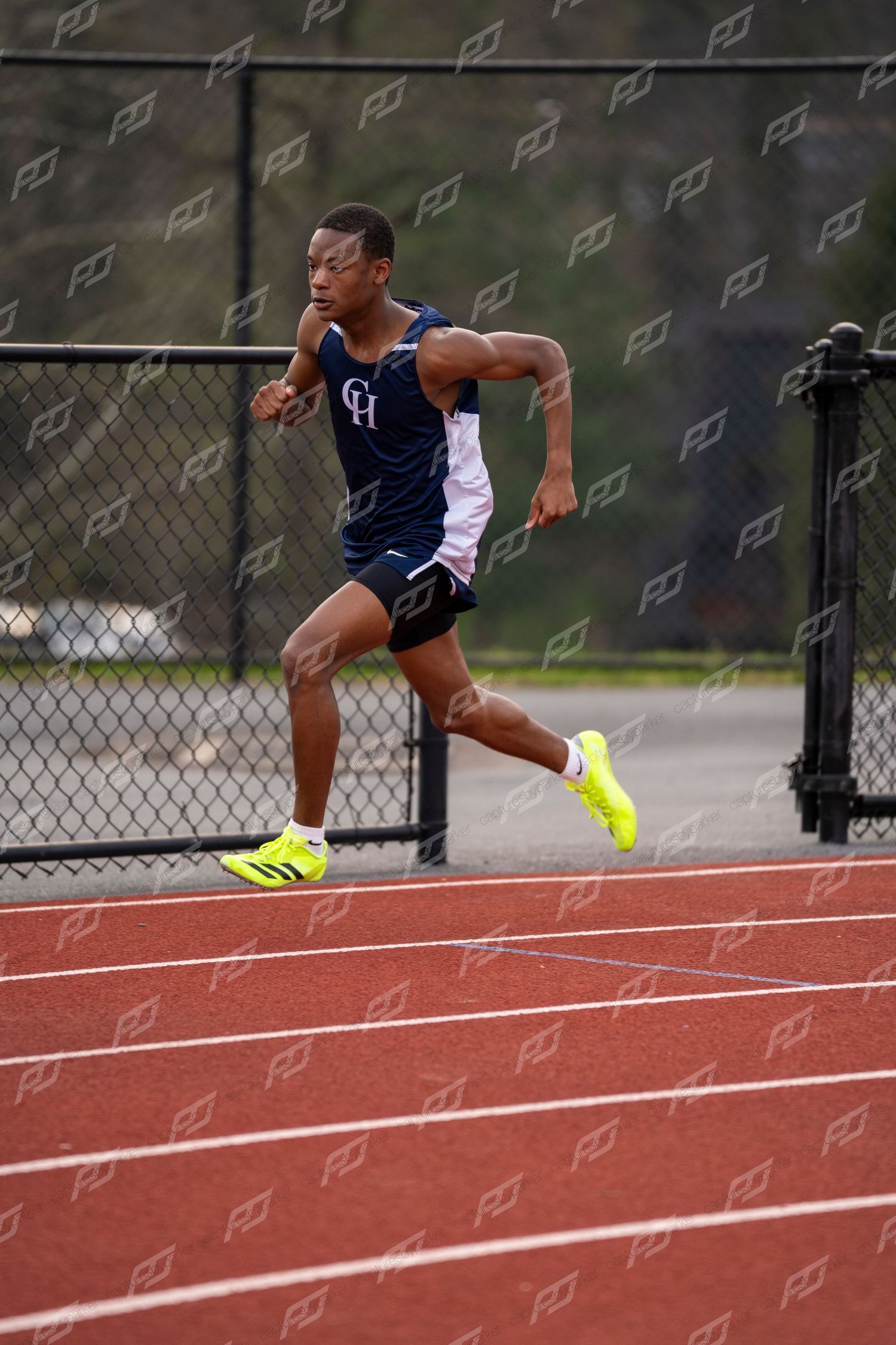 Male sprinter in navy CH uniform runs powerfully on red track wearing neon yellow shoes.