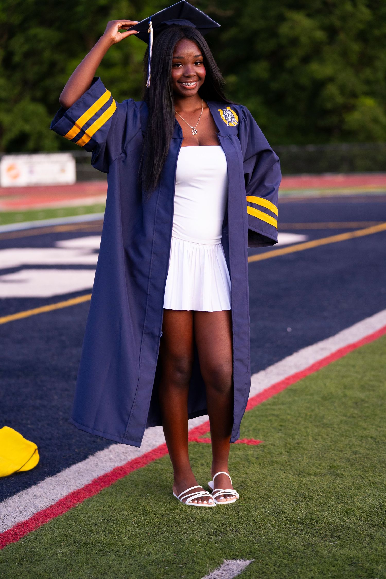 Smiling female graduate adjusts navy cap and gown with gold trim on athletic track field.