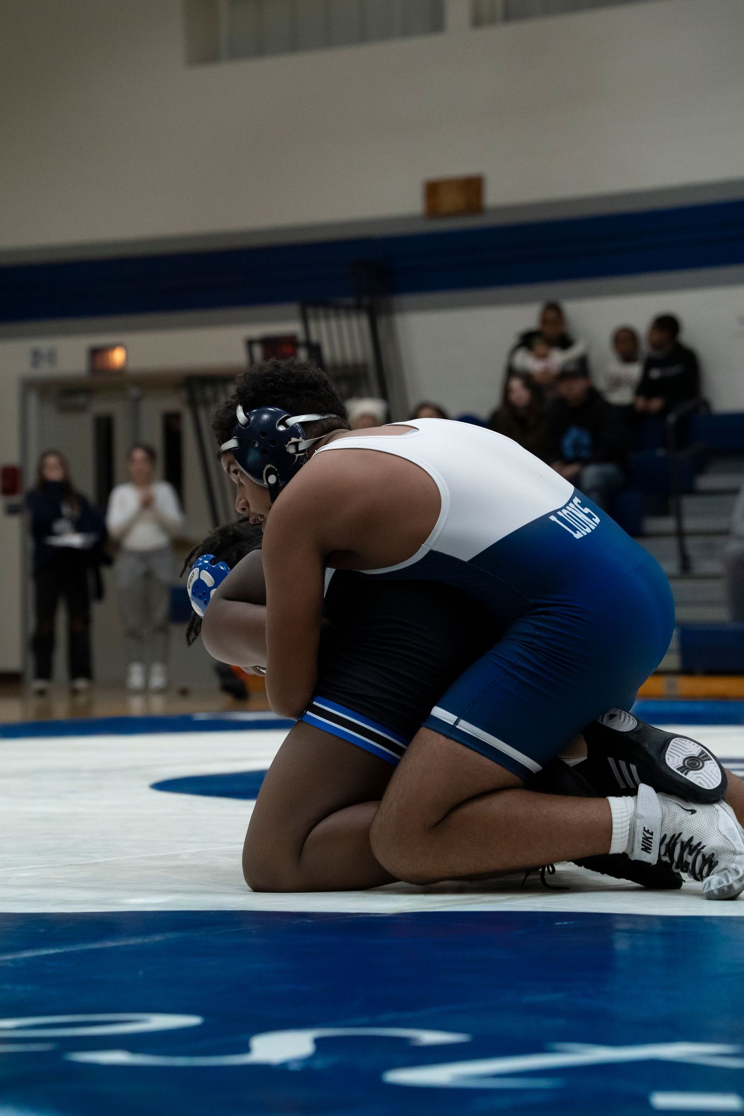 Wrestler in white and blue singlet controls opponent on knees during competitive high school wrestling match.