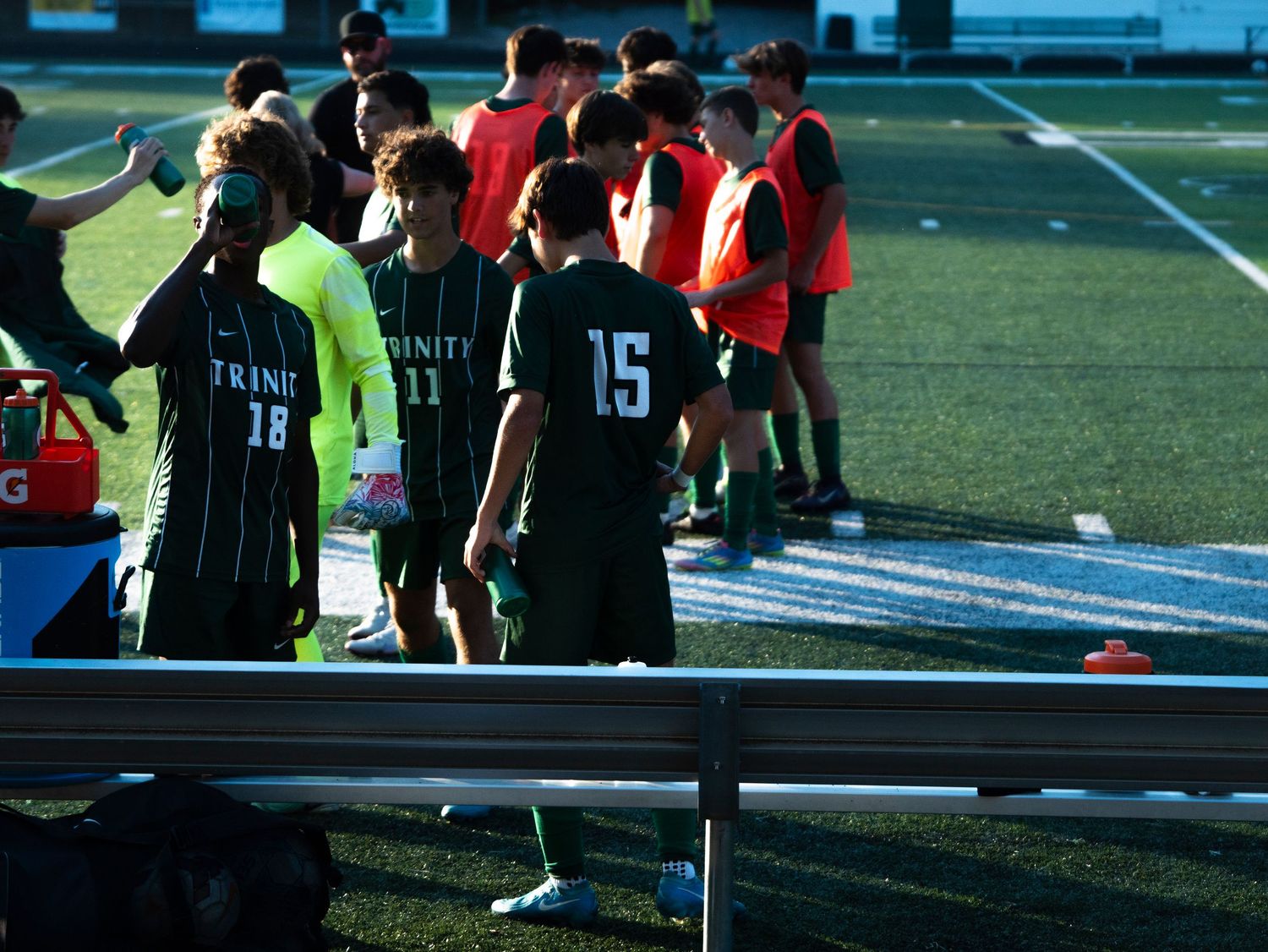 Trinity soccer players #18 and #15 greet teammates near sideline bench during daytime match on turf.