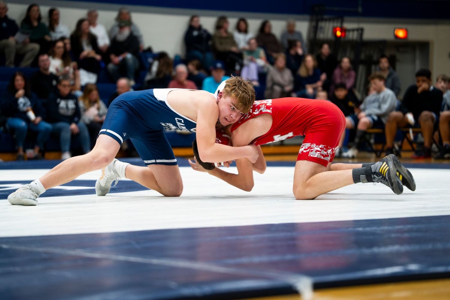 Two high school wrestlers grapple on the mat, one in blue and one in red singlet.