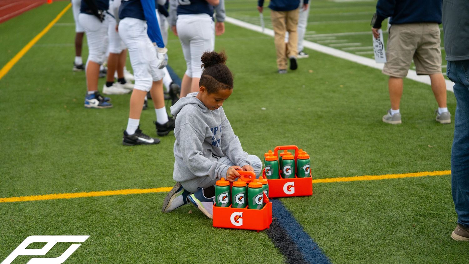 Young child organizes Gatorade bottles on a turf field sideline during a youth football game.