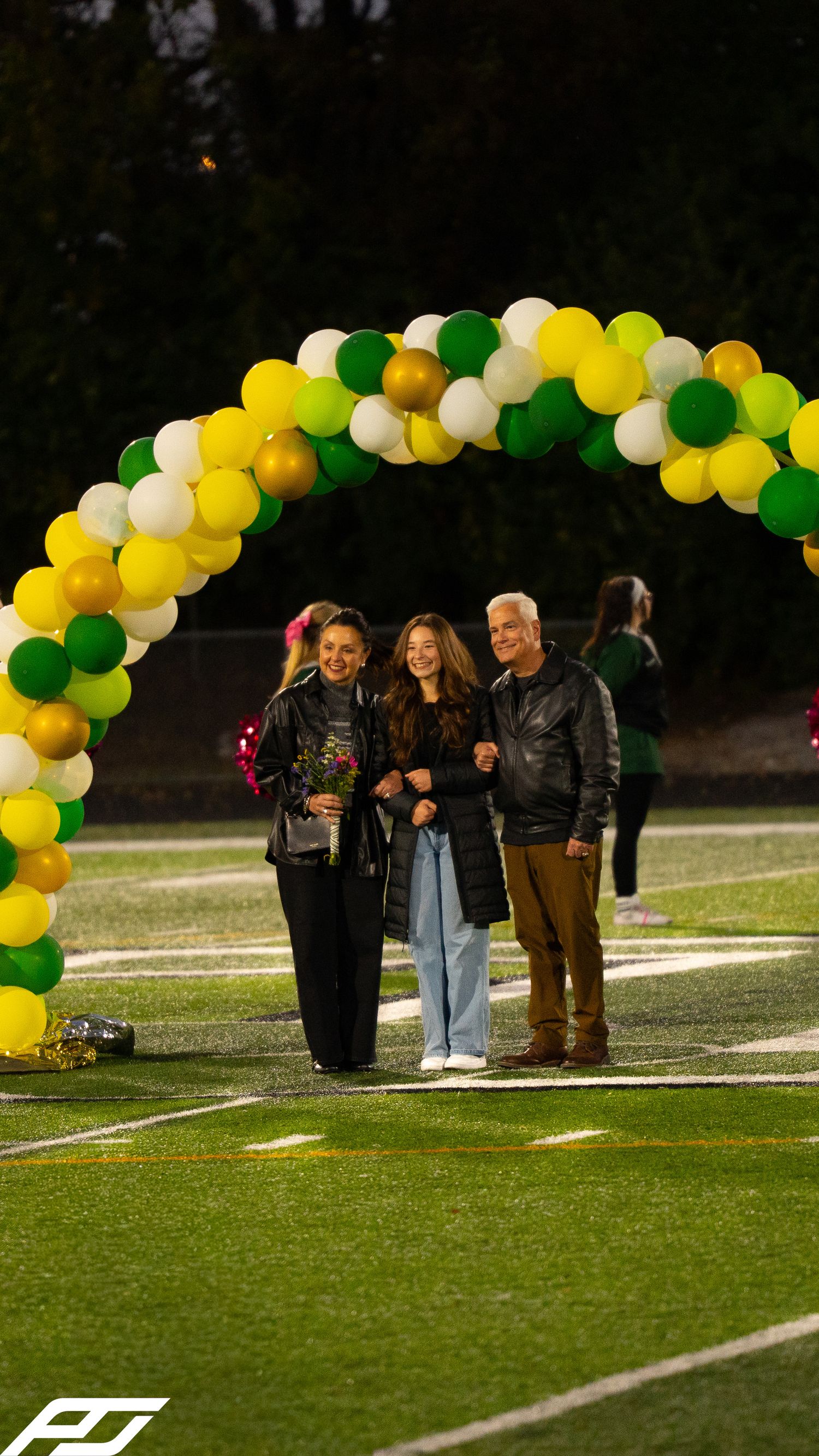 Senior student celebrates with parents under green, gold, white balloon arch during evening football field ceremony.