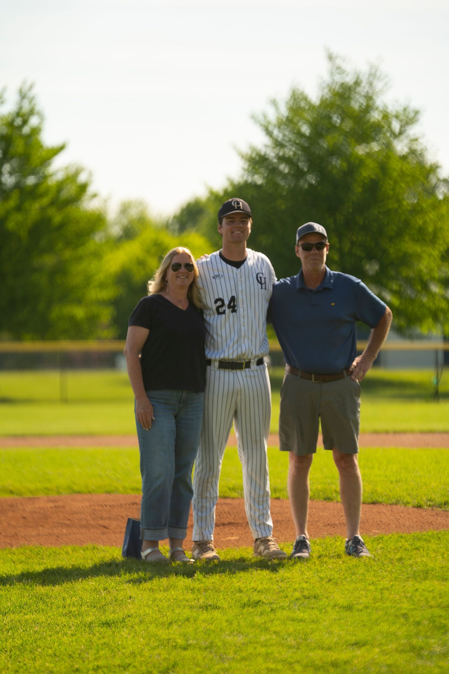 Baseball player #24 in pinstripe uniform smiles with two parents on a bright sunny baseball field.