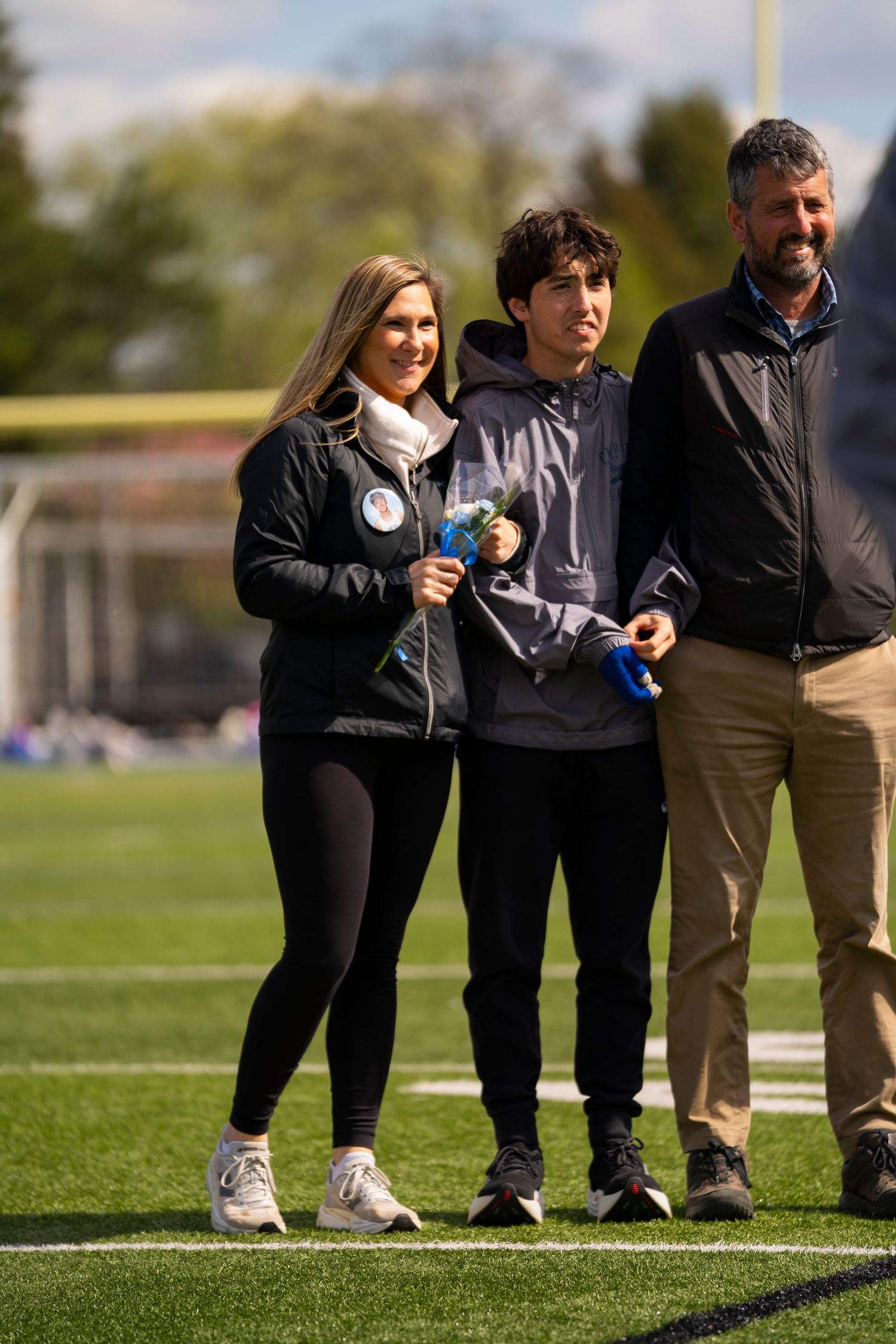 A young athlete holds trophies while posing with two smiling adults on a green sports field.