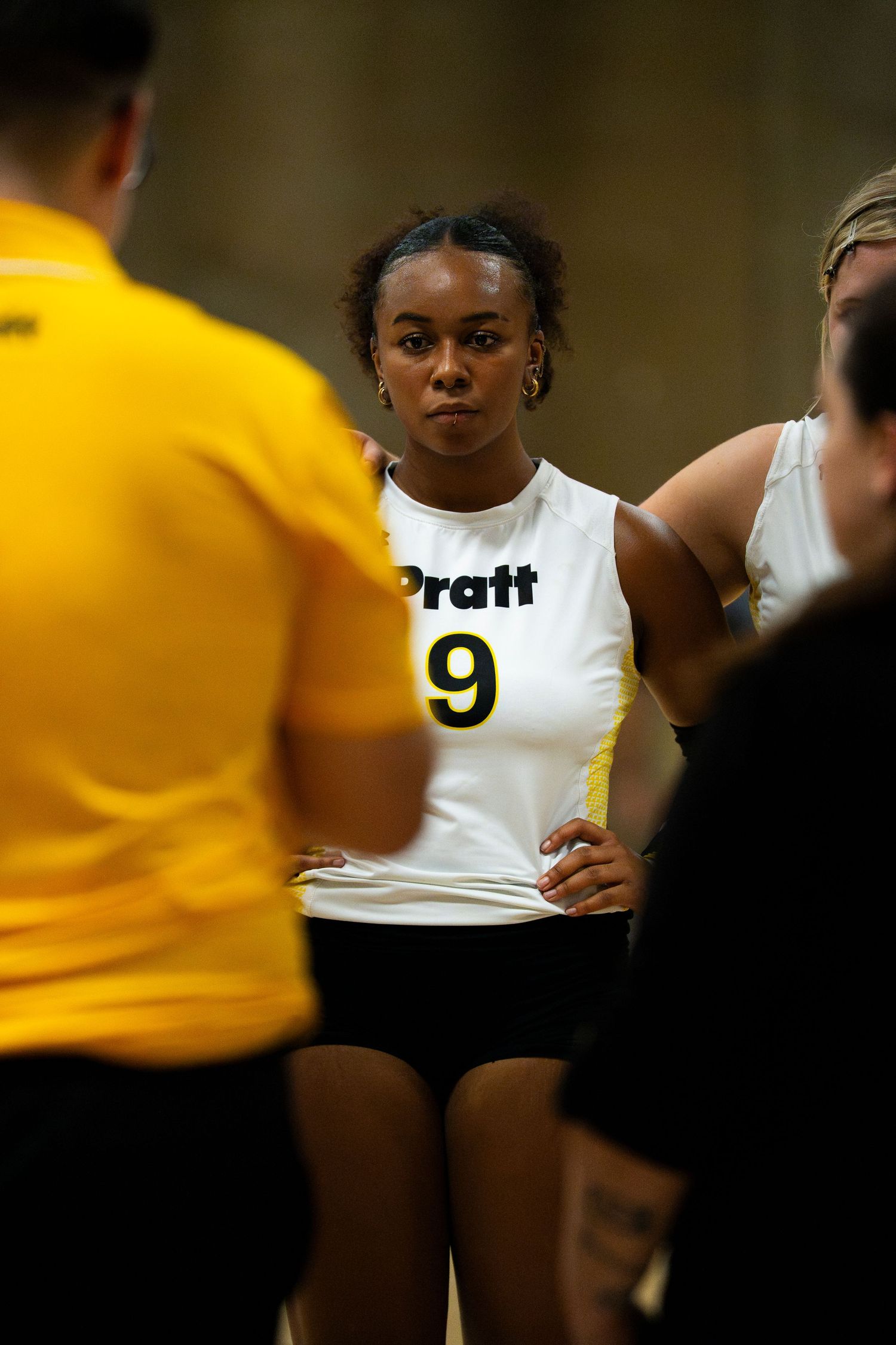 Pratt volleyball player #9 listens intently during a team huddle wearing a white jersey.