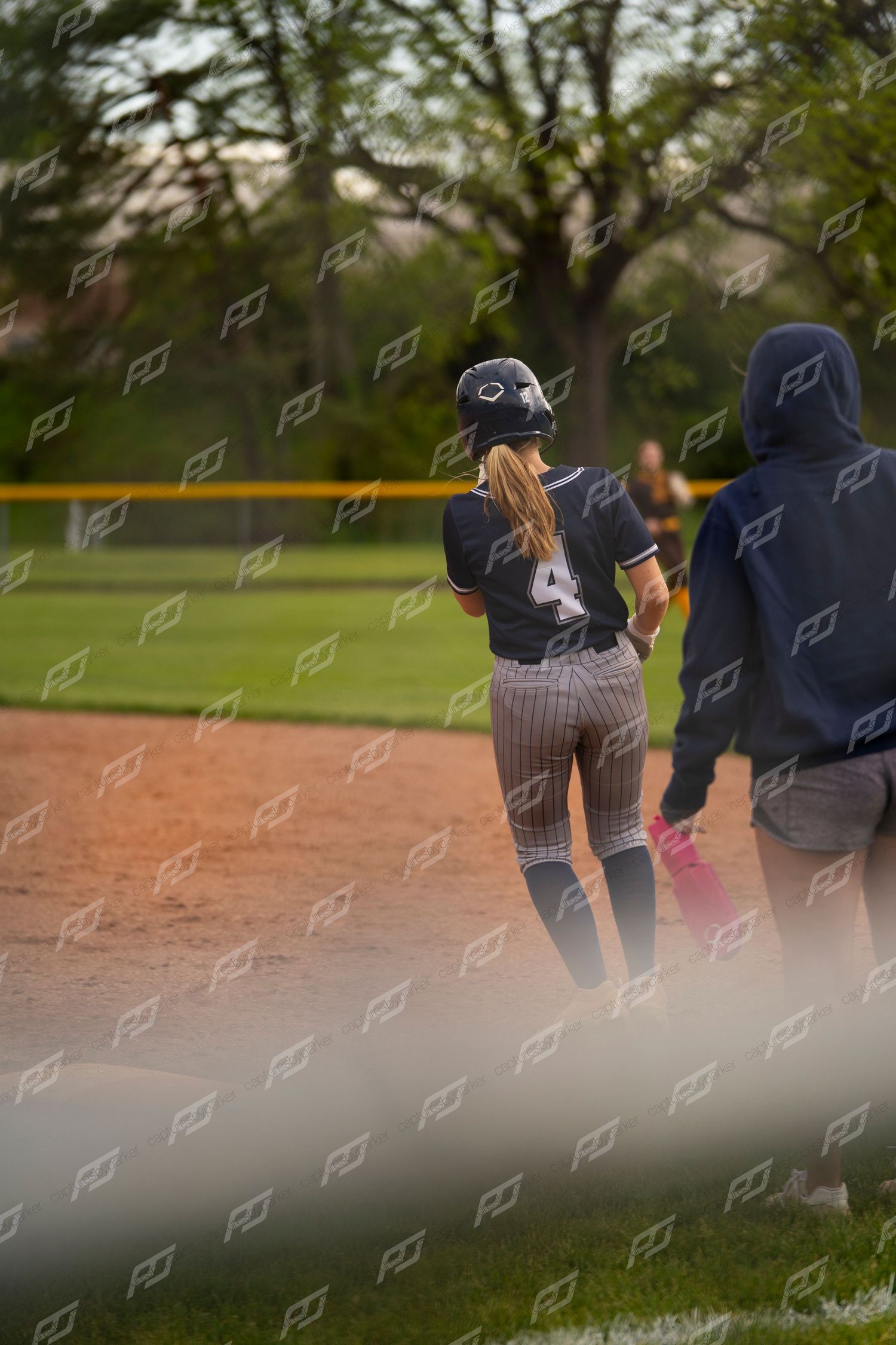 Softball player wearing number 4 stands near a base with a coach during a game.