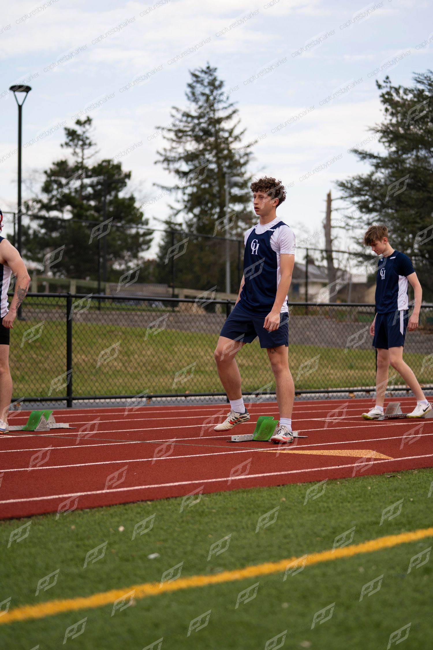 Three male track athletes in navy uniforms stand near starting blocks on red track.