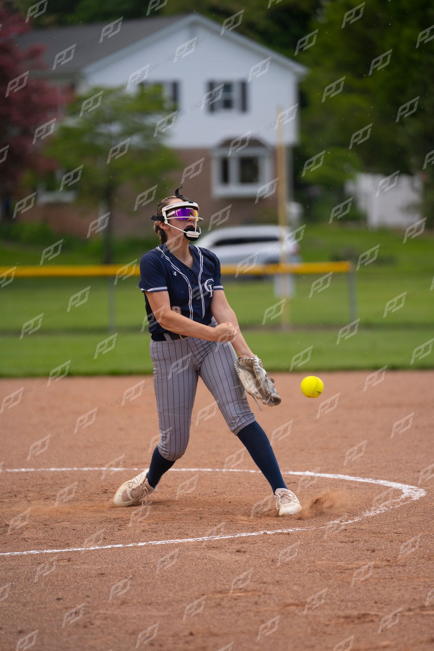 Female softball pitcher in navy uniform winds up to deliver an underhand pitch on a dirt mound.