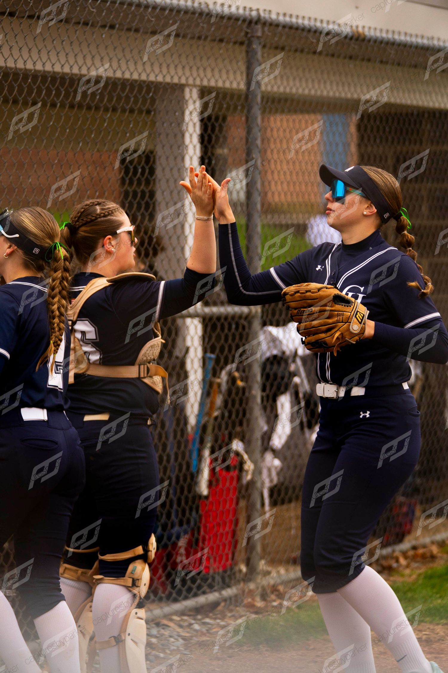 Softball teammates in navy uniforms exchanging high fives near the dugout chain-link fence during game.