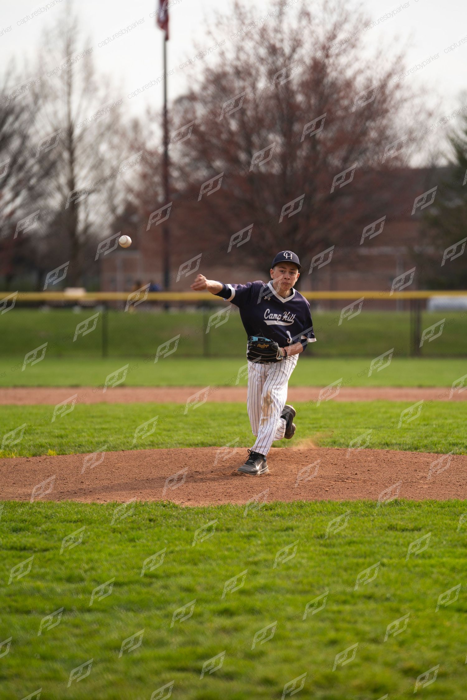 Young baseball pitcher in navy pinstripe uniform releasing a pitch from the mound during a spring game.