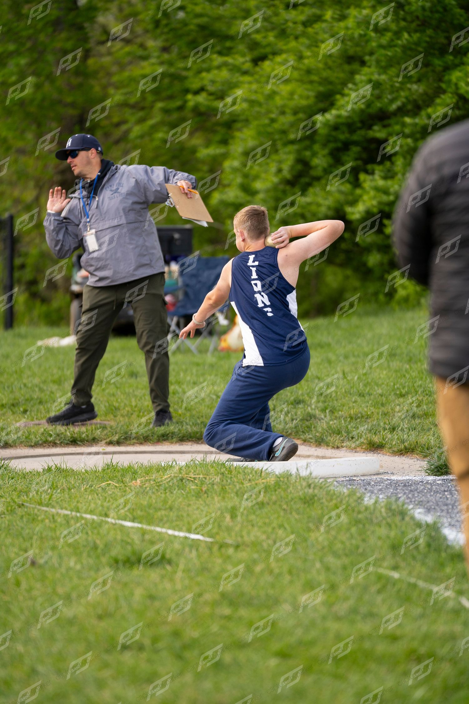 Male LIONS athlete throwing discus while official signals at outdoor track and field competition.