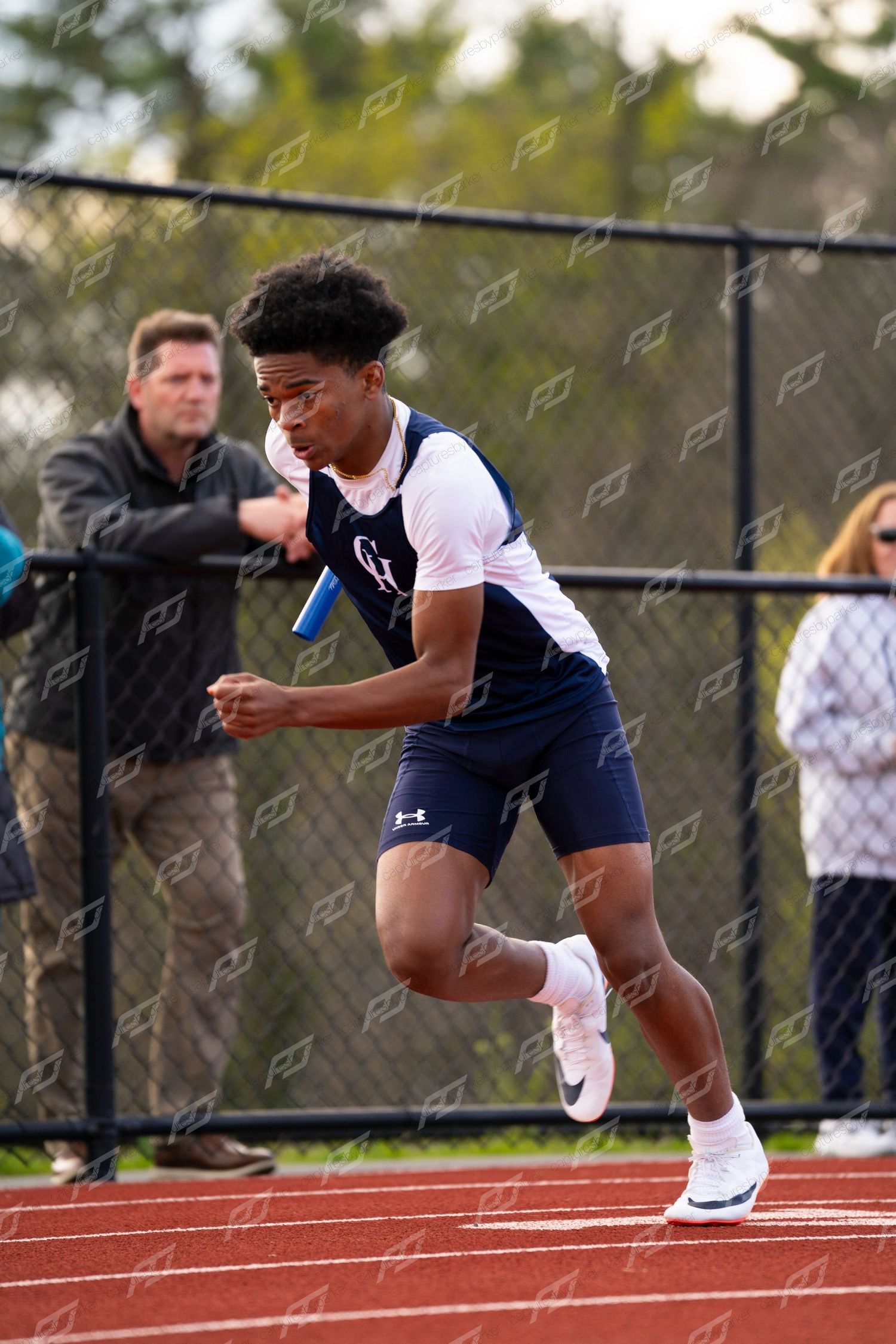 Male high school track athlete accelerates from relay exchange zone gripping blue baton during outdoor track meet.