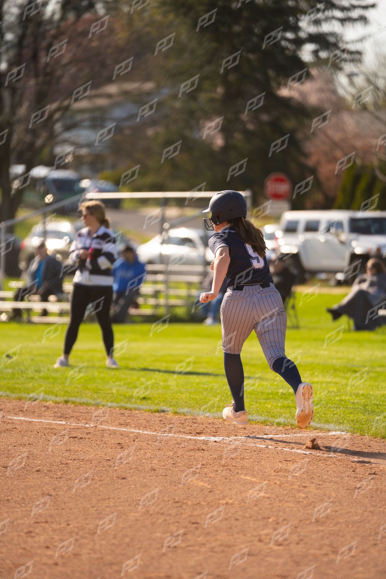 Female softball player #5 in navy uniform running bases on dirt infield during outdoor game.