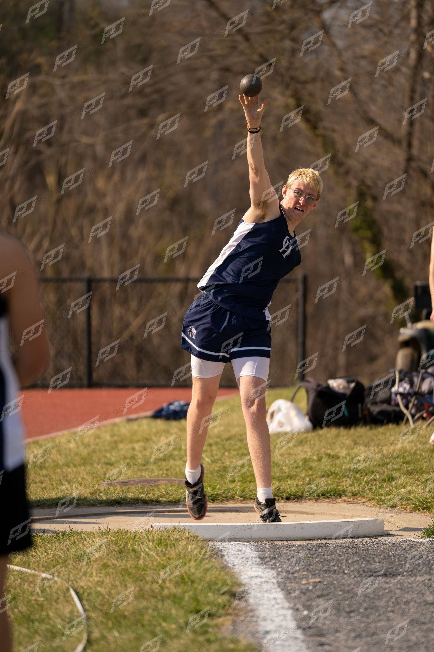 Young male athlete throwing shot put with arm fully extended at outdoor track meet.