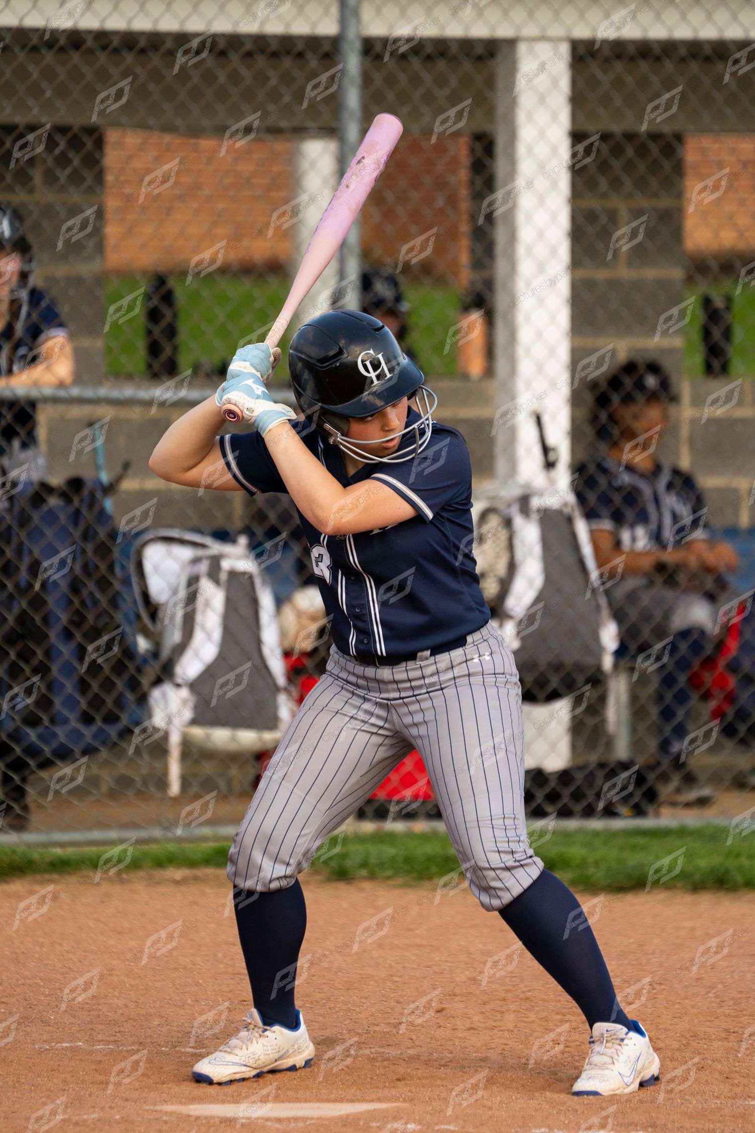 Female softball batter in navy pinstripe uniform stands ready at plate holding pink bat.