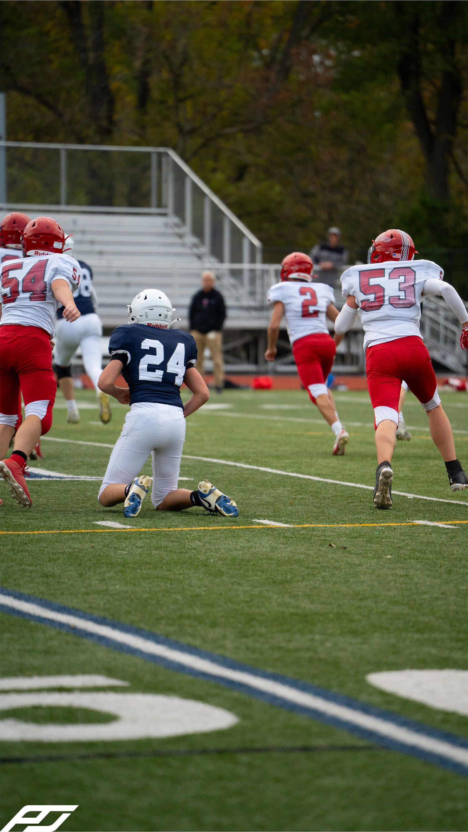 Youth football player number 24 in navy kneels on turf as red-uniformed opponents run downfield.