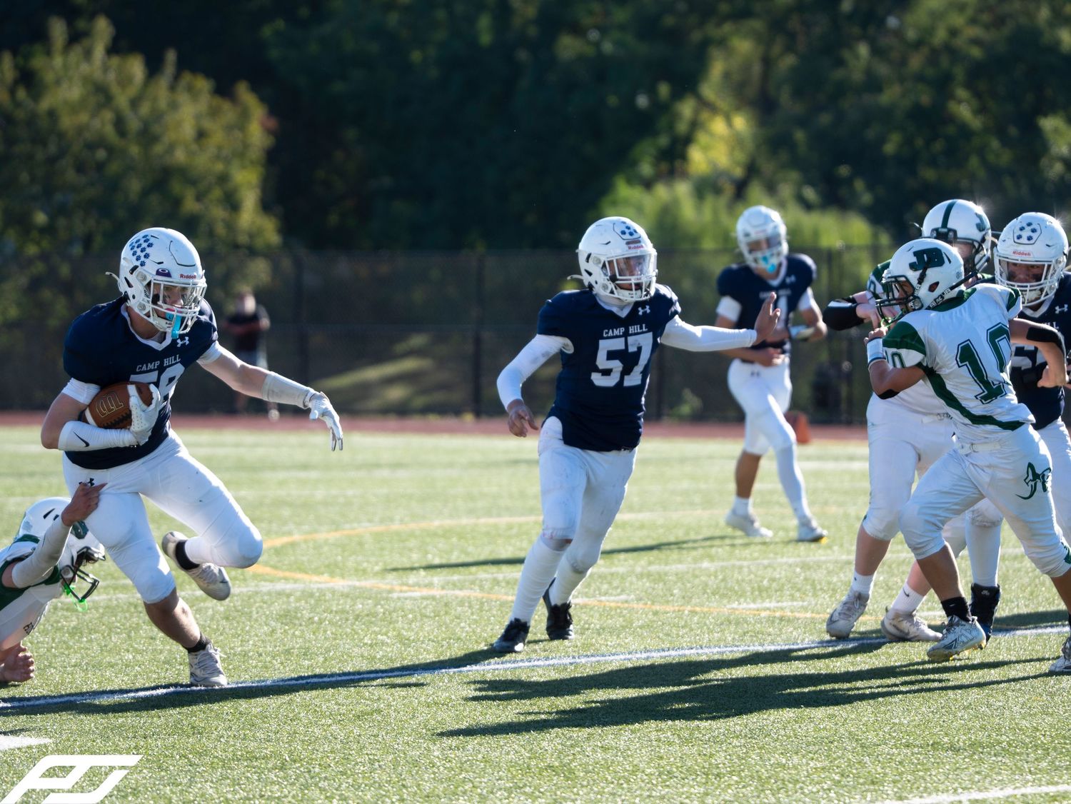 Youth football players in navy uniforms compete during a game, with player 57 blocking on a sunny field.