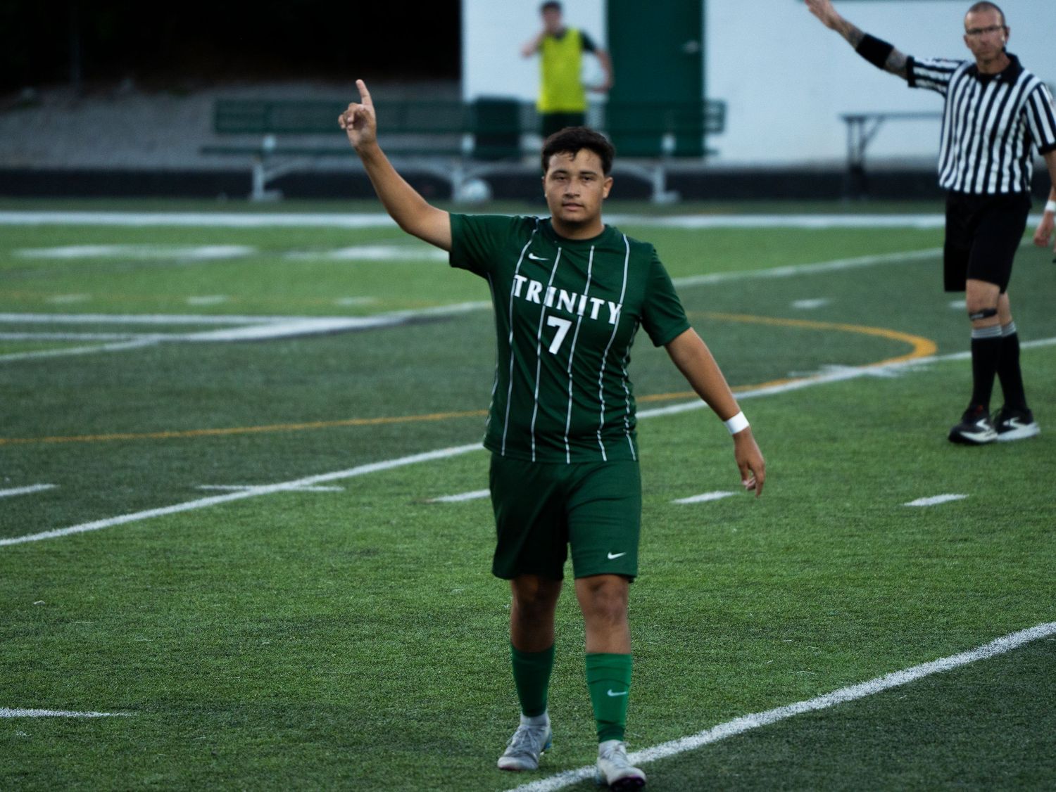 Trinity soccer player #7 raises finger in celebration on turf field with referee signaling in background.