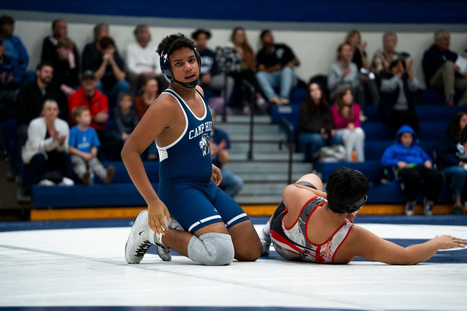 High school wrestler in blue singlet stands over defeated opponent on wrestling mat after winning match.