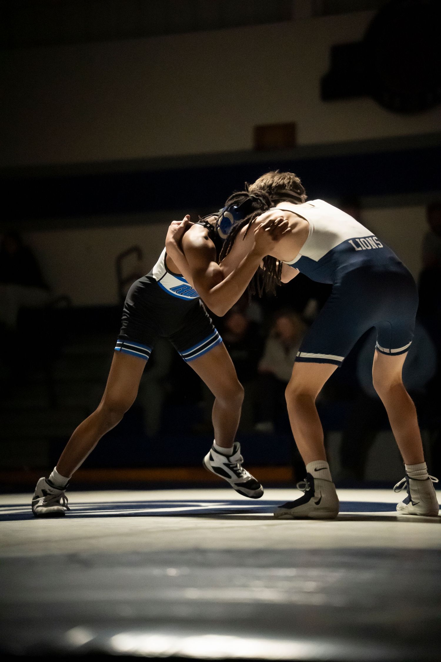 Two wrestlers in blue and white singlets grapple standing during a competitive high school wrestling match.