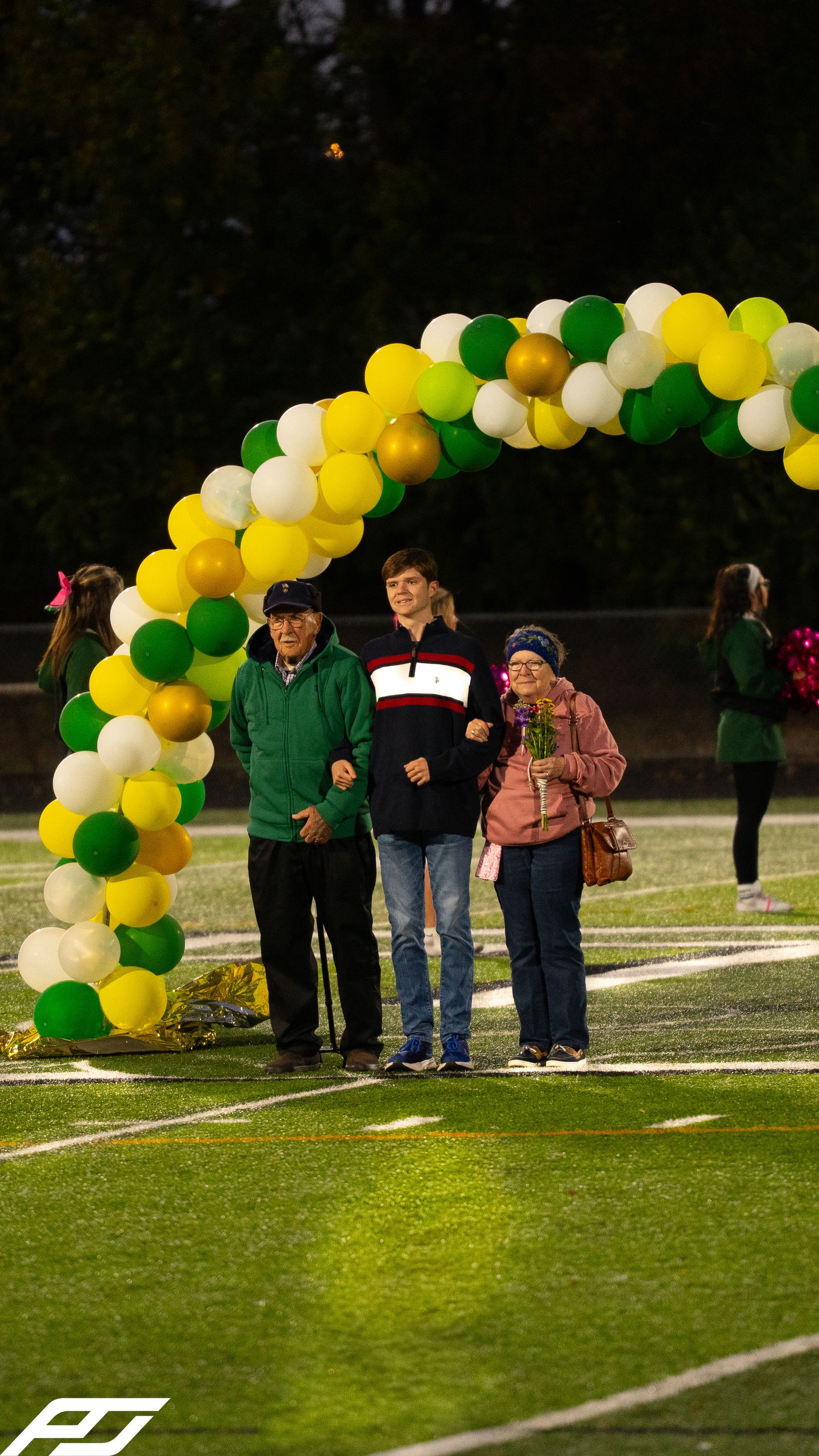 Senior student walks with grandparents under green and gold balloon arch at evening football field ceremony.