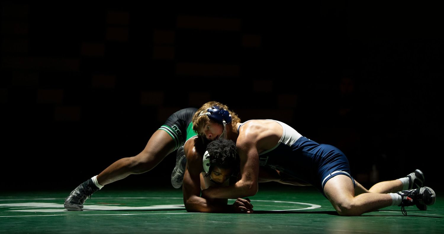 Two wrestlers compete in low grappling position on mat under dramatic dark lighting.