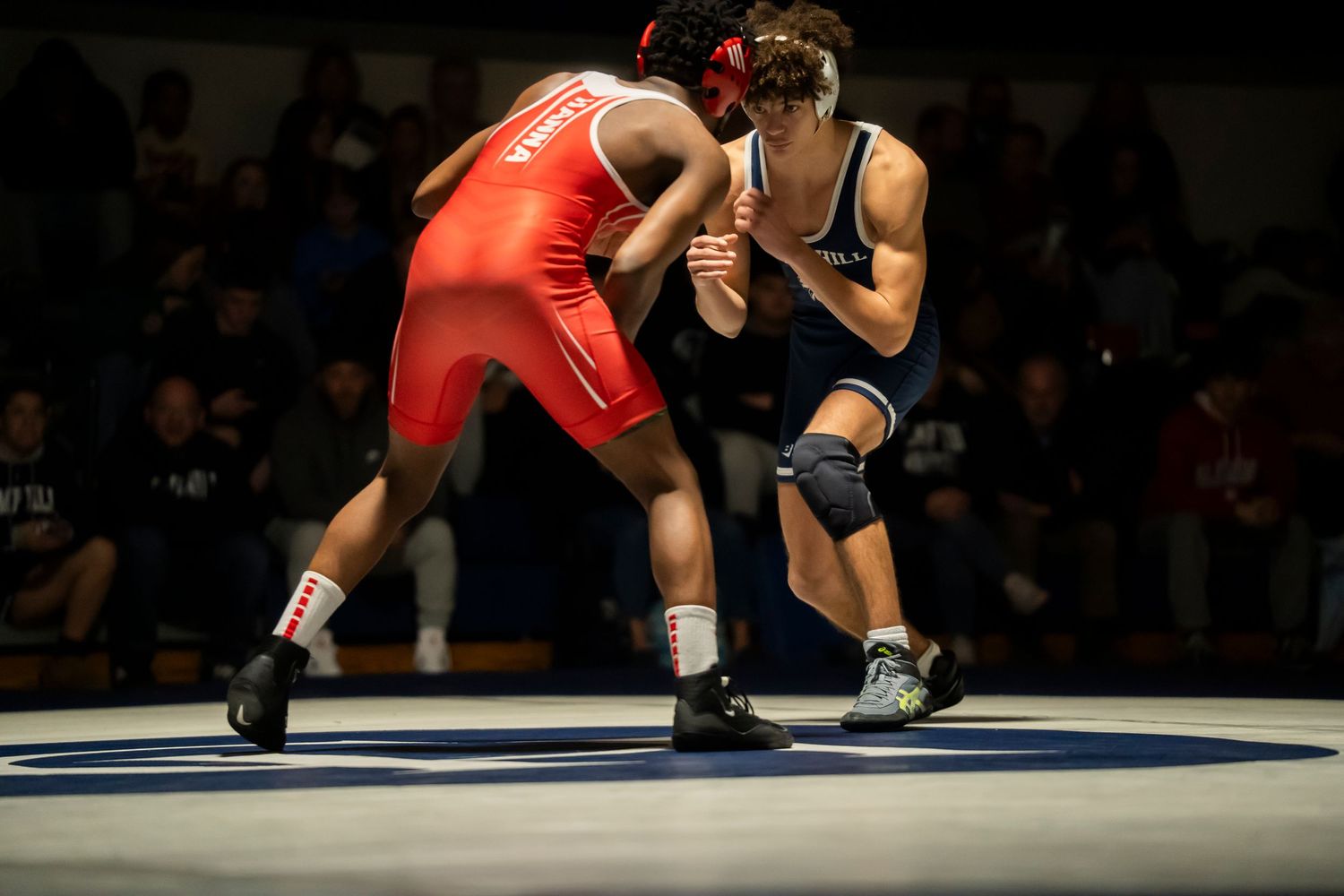Two wrestlers in red and blue singlets engage in a standing grapple during a wrestling competition.