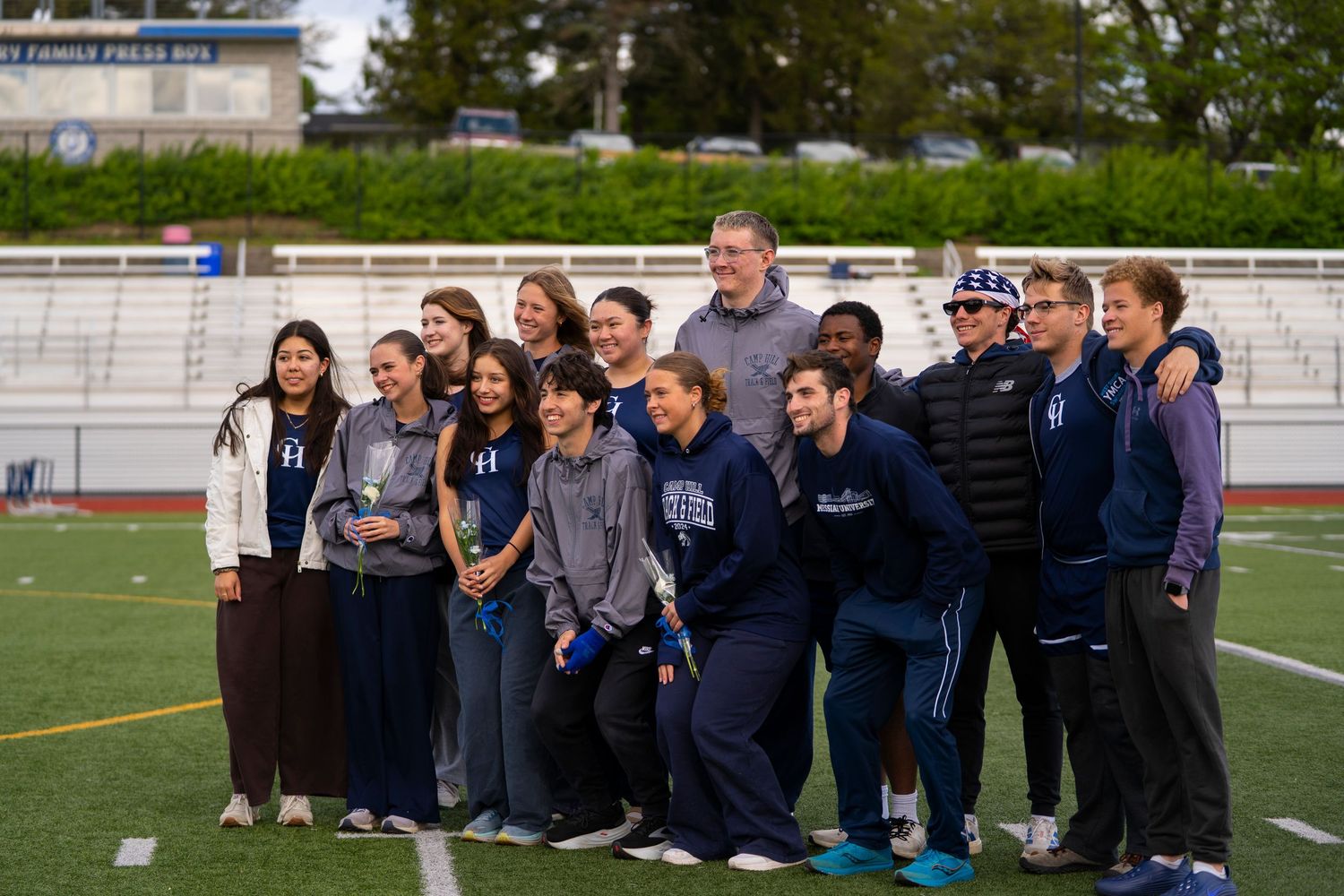 Large group of students and adults pose together on a football field wearing navy and gray team apparel.