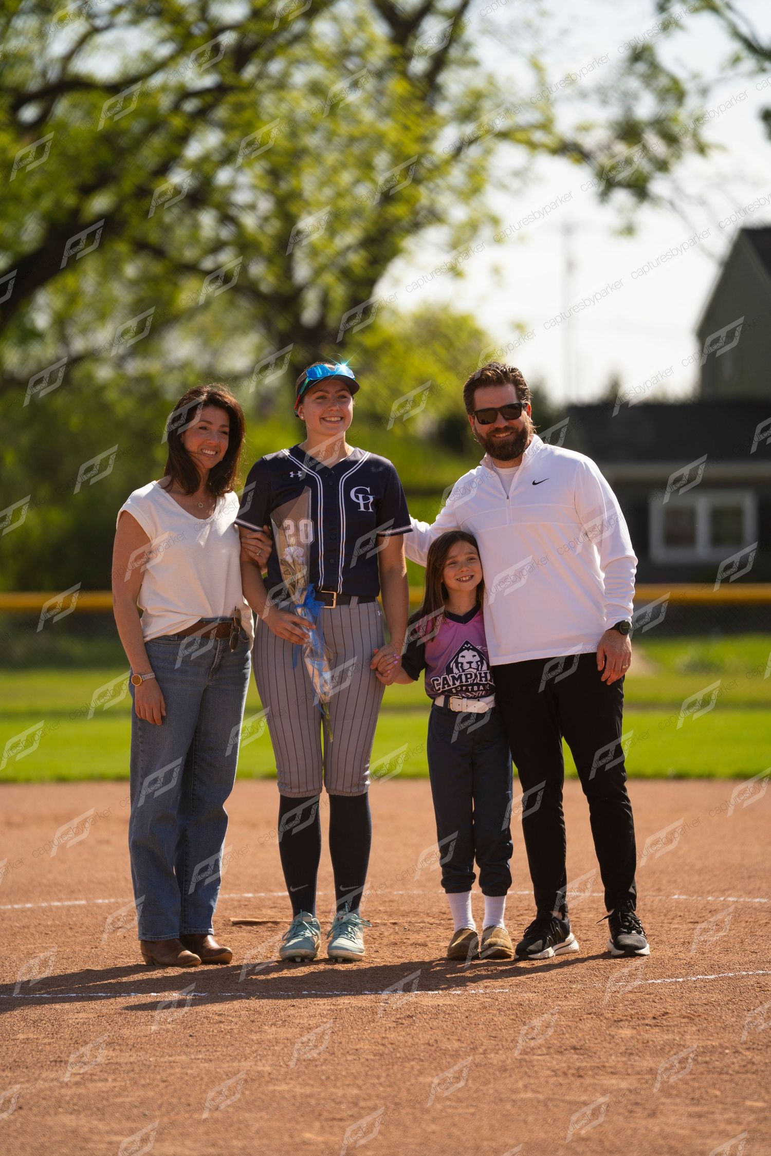Softball player in navy uniform poses with family of four on baseball diamond on sunny day.