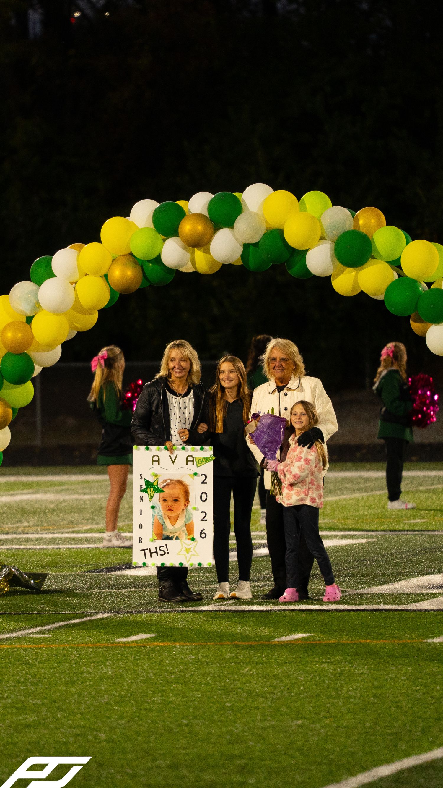 Family celebrates Ava 2026 with a poster and flowers under a green and yellow balloon arch at senior night.